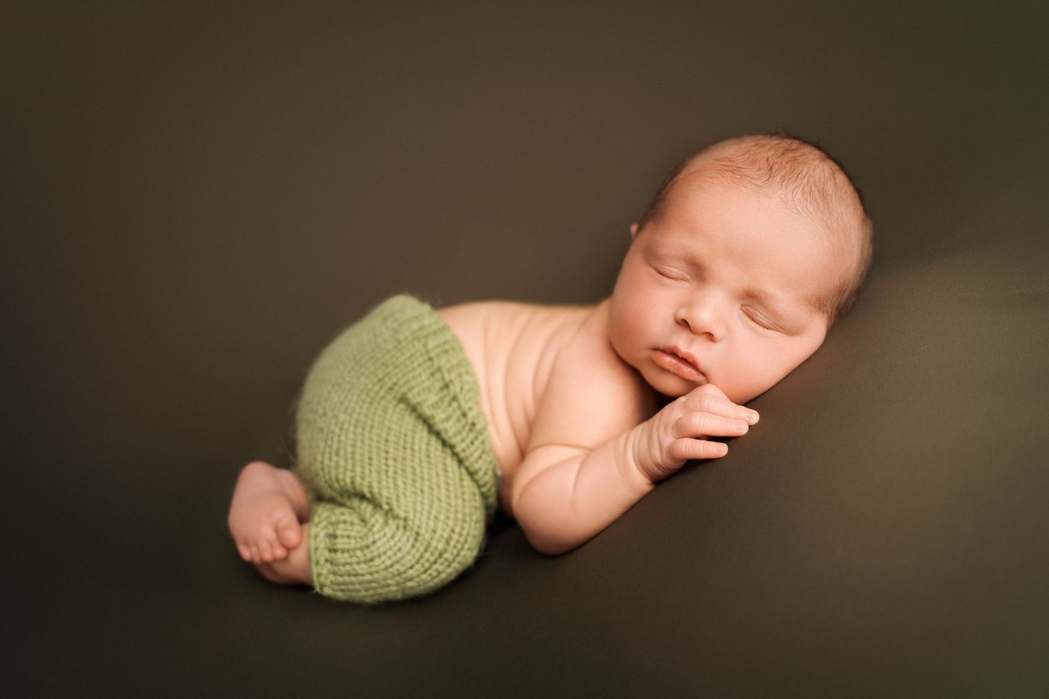 Peaceful newborn baby sleeping on a green backdrop wearing sage green knit pants.