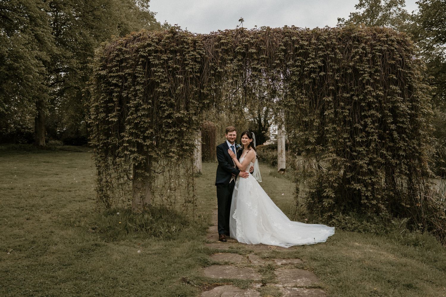 Bride and groom embrace beneath a dramatic ivy-covered archway in a lush garden on their wedding day.