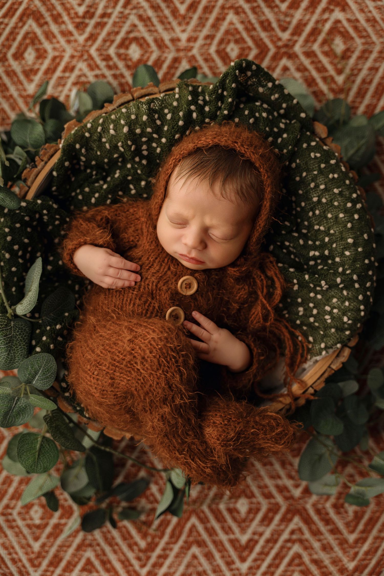 Sleeping newborn baby in brown knit outfit rests in a basket with green eucalyptus leaves.