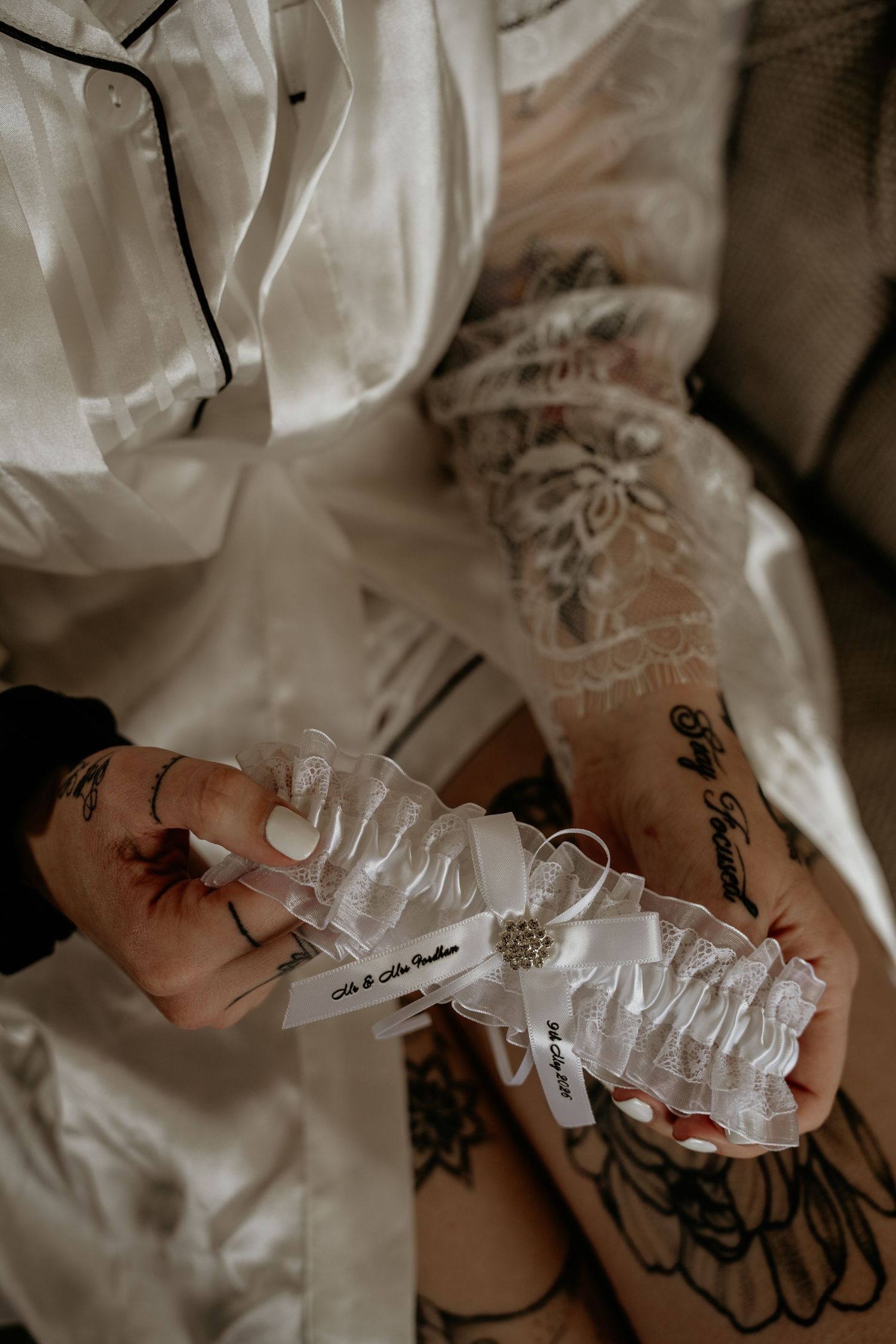 Tattooed bride in a silk robe holds a personalized white lace wedding garter in her hands.