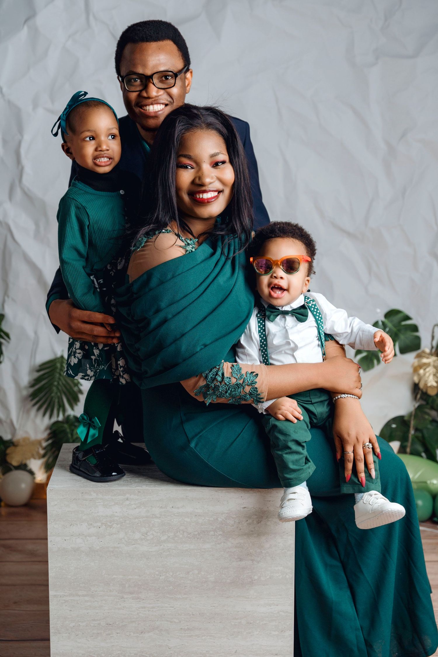 A joyful Black family of four poses in coordinated green outfits, with two young children, in a bright studio setting.