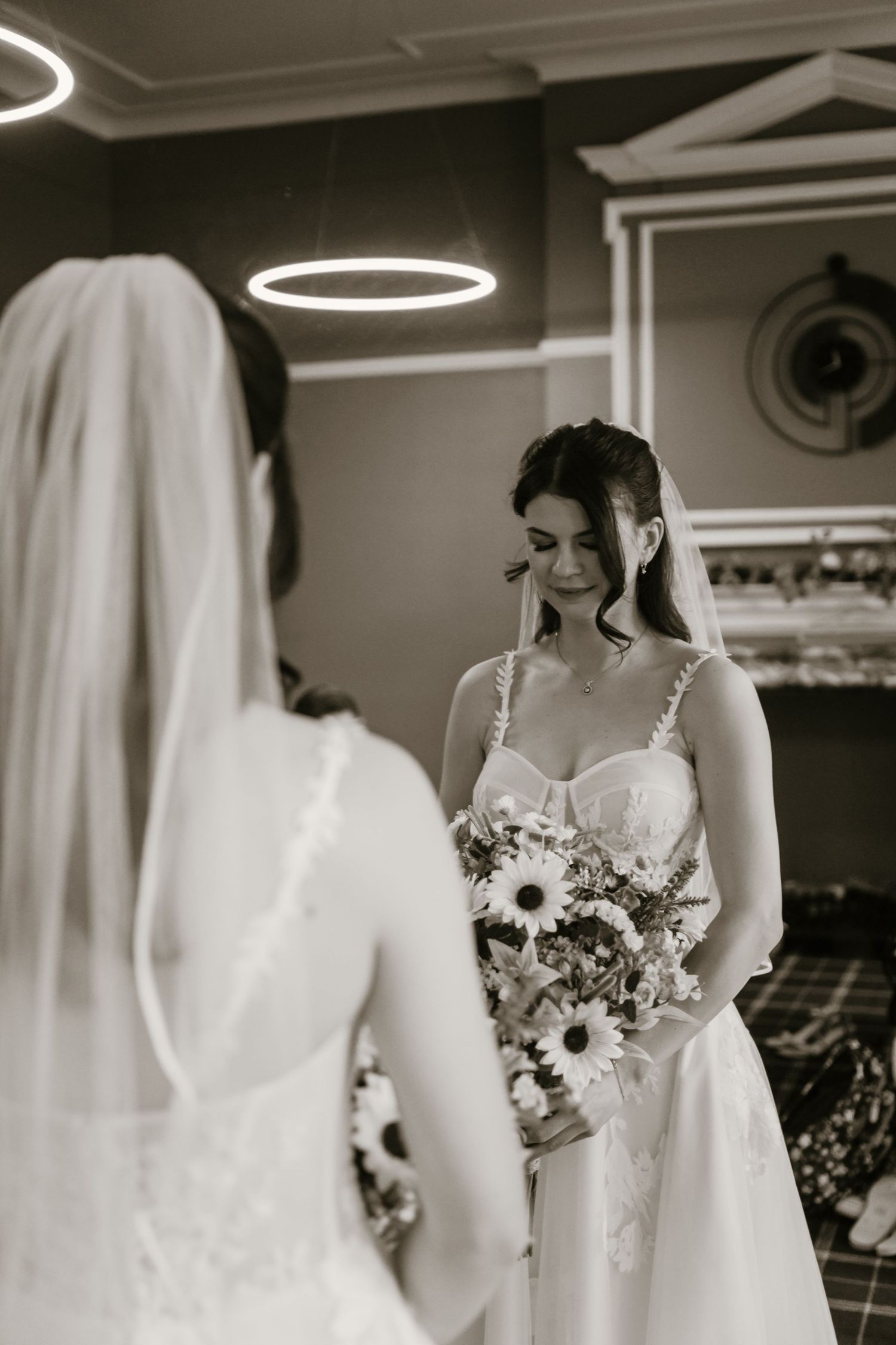 Bride in white gown holding sunflower bouquet gazes down in black and white mirror reflection photo.