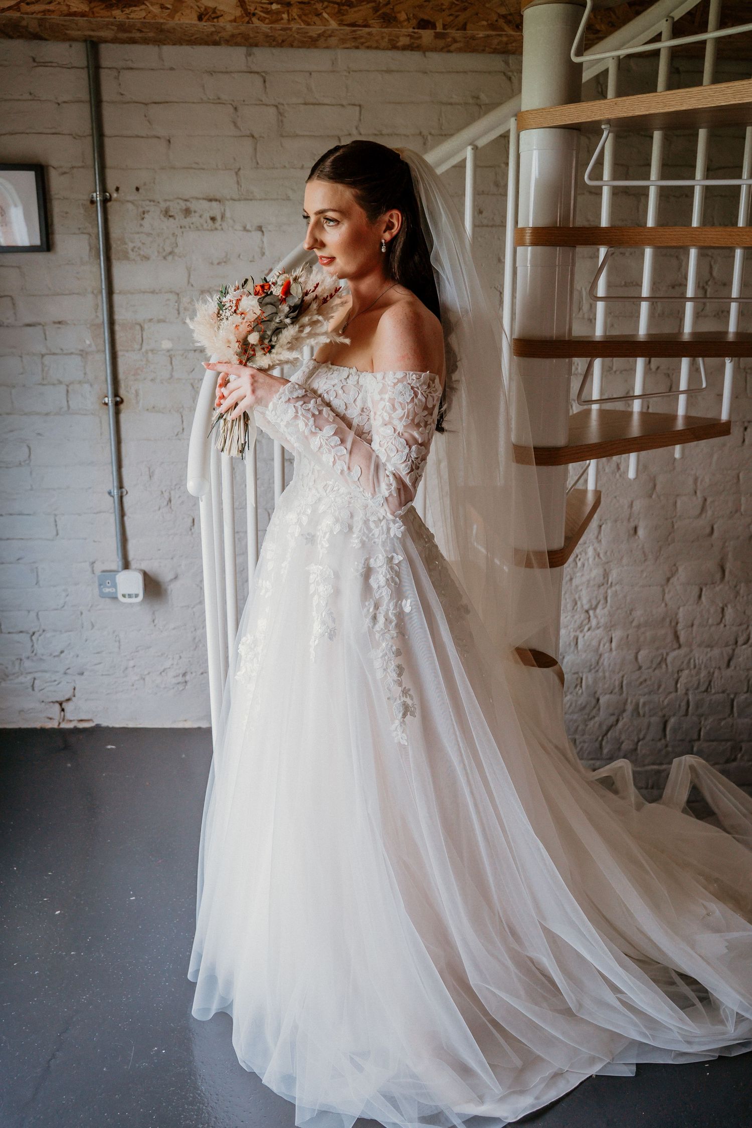 Bride in off-shoulder lace wedding gown holding floral bouquet near spiral staircase in white brick room.