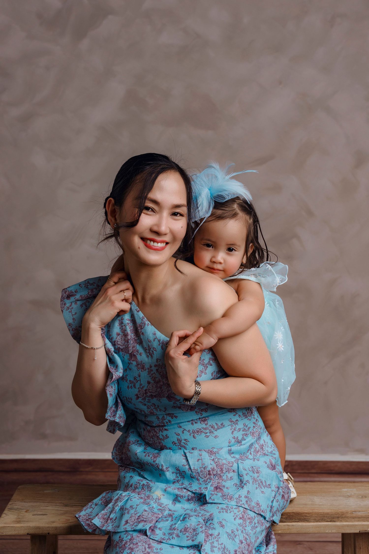 Smiling Asian woman in blue floral dress holding baby girl in matching blue outfit with feather headband.