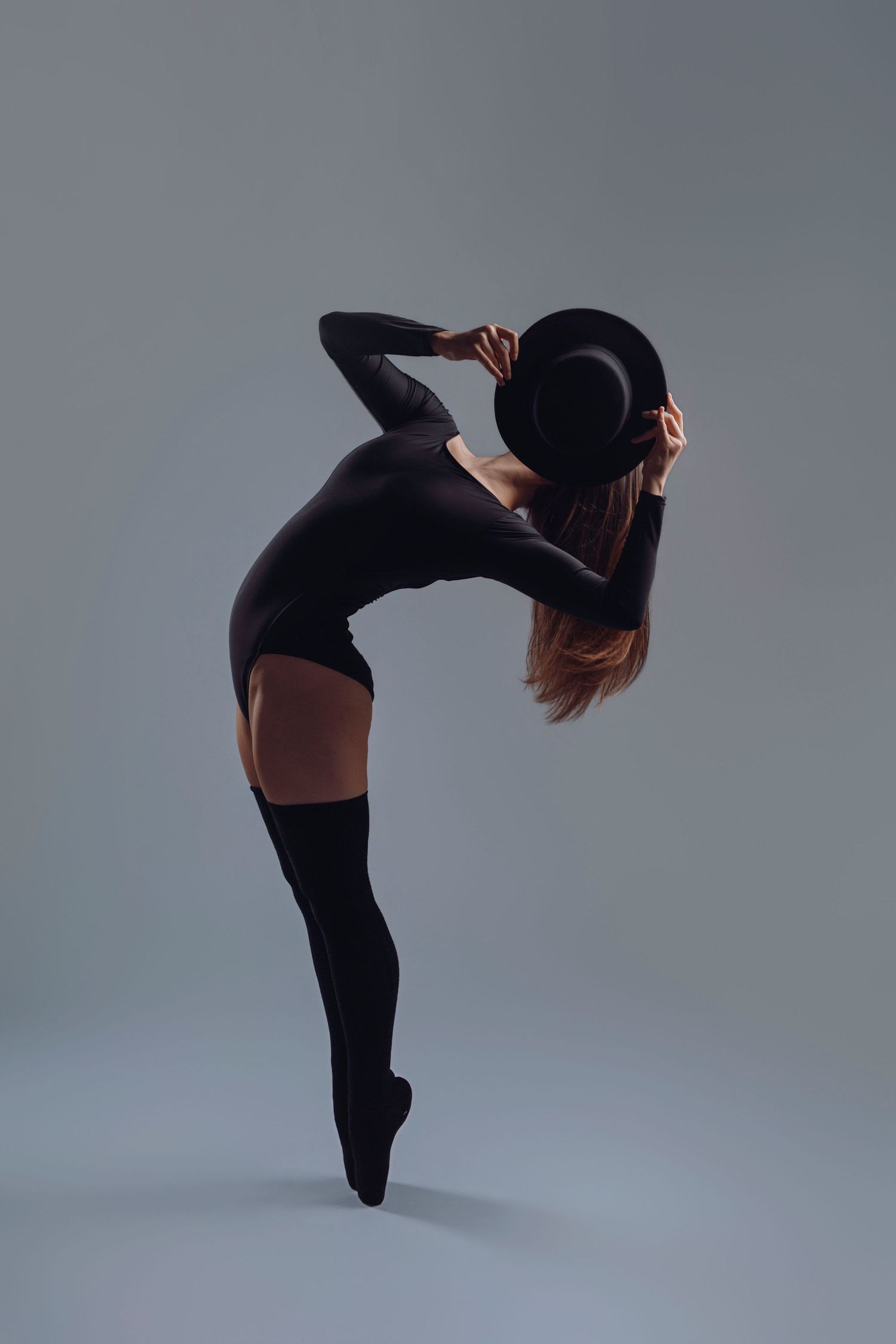 Elegant dancer in black bodysuit and hat striking a graceful backbend pose on pointe against gray background.
