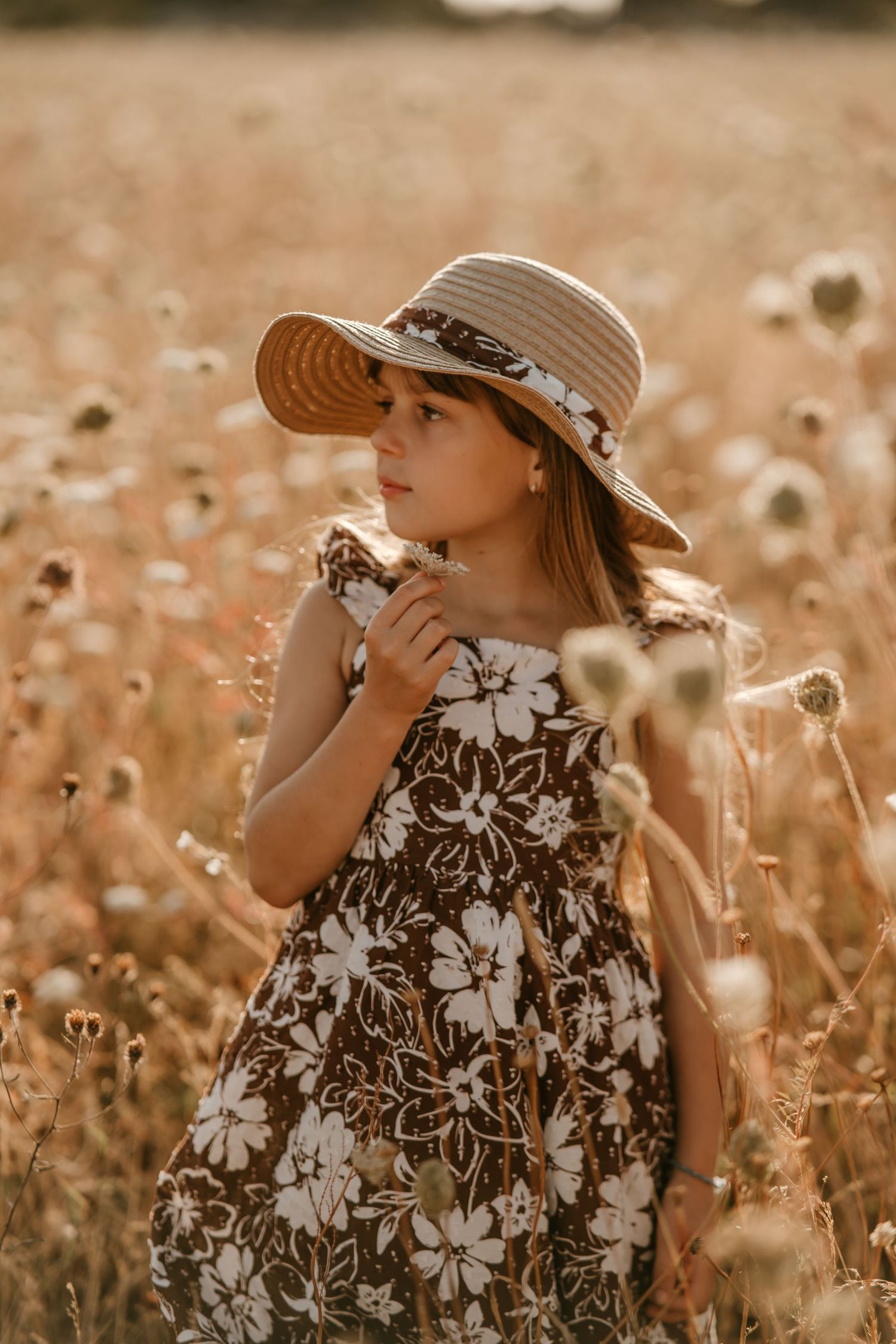 Young girl in floral dress and straw hat standing in golden field holding wildflowers at sunset.