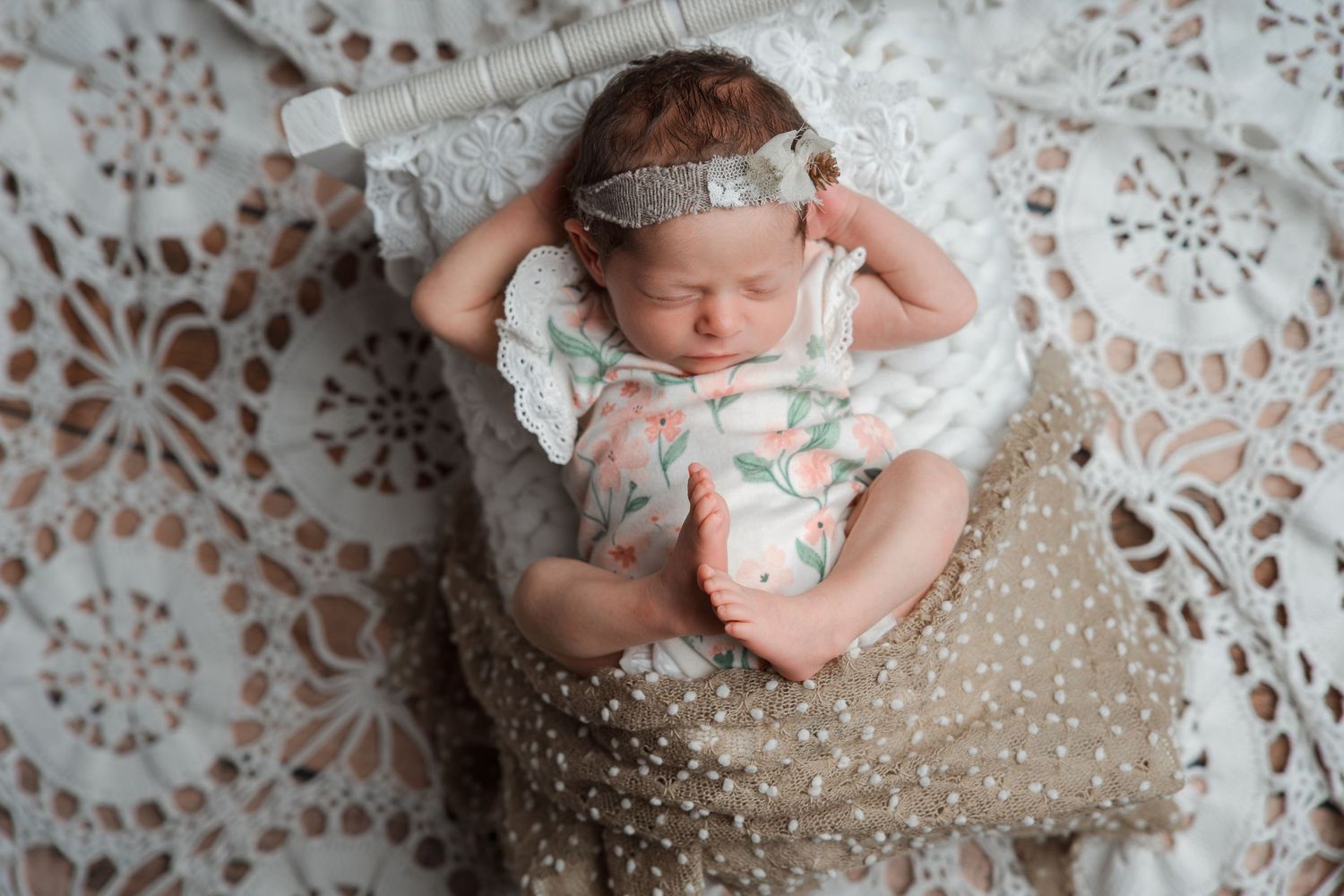 Newborn baby girl sleeping in floral romper with lace headband on white crochet blanket and polka dot pillow.
