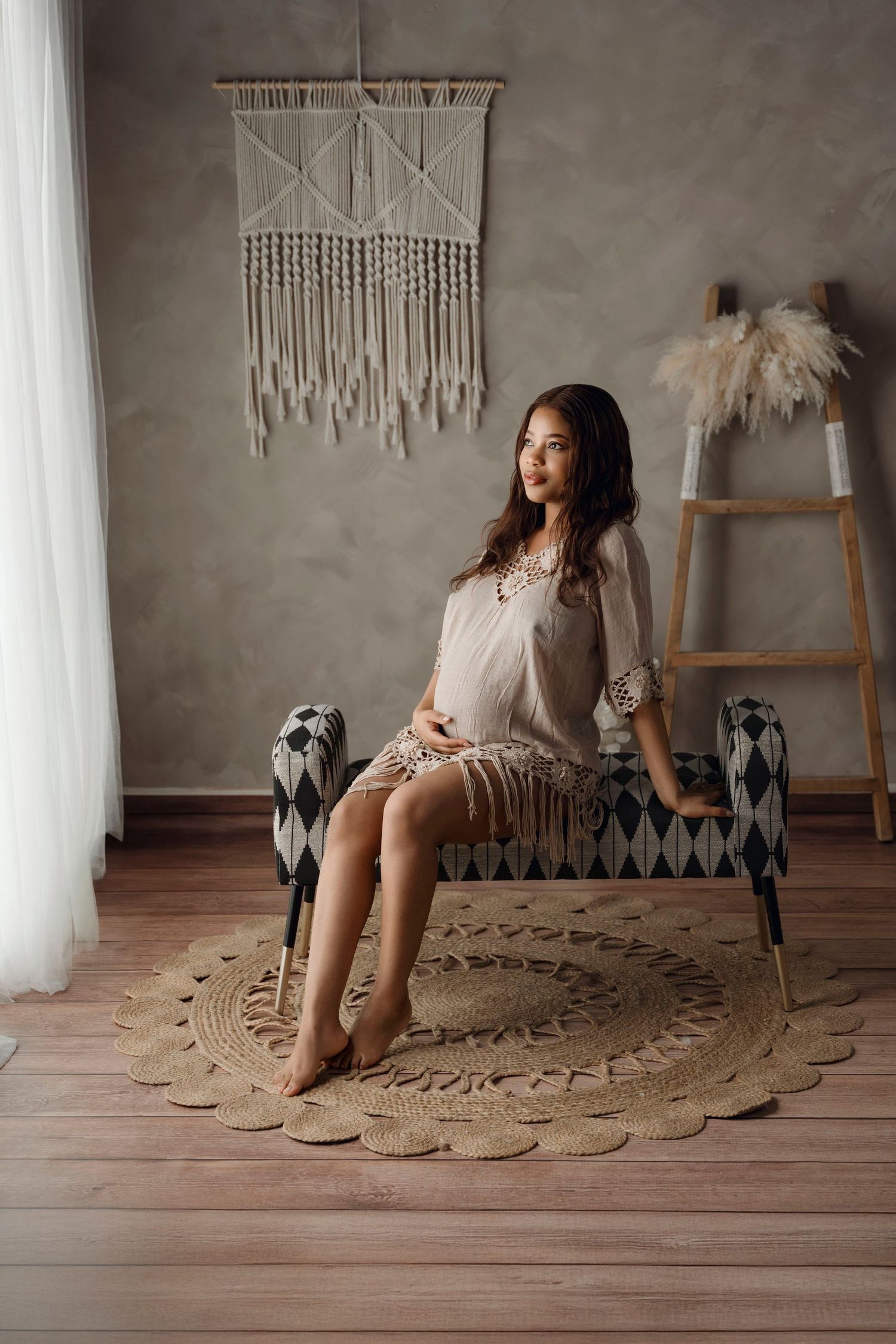 Pregnant woman in a beige fringe dress sitting on a bench in a boho-styled room with macrame wall decor.
