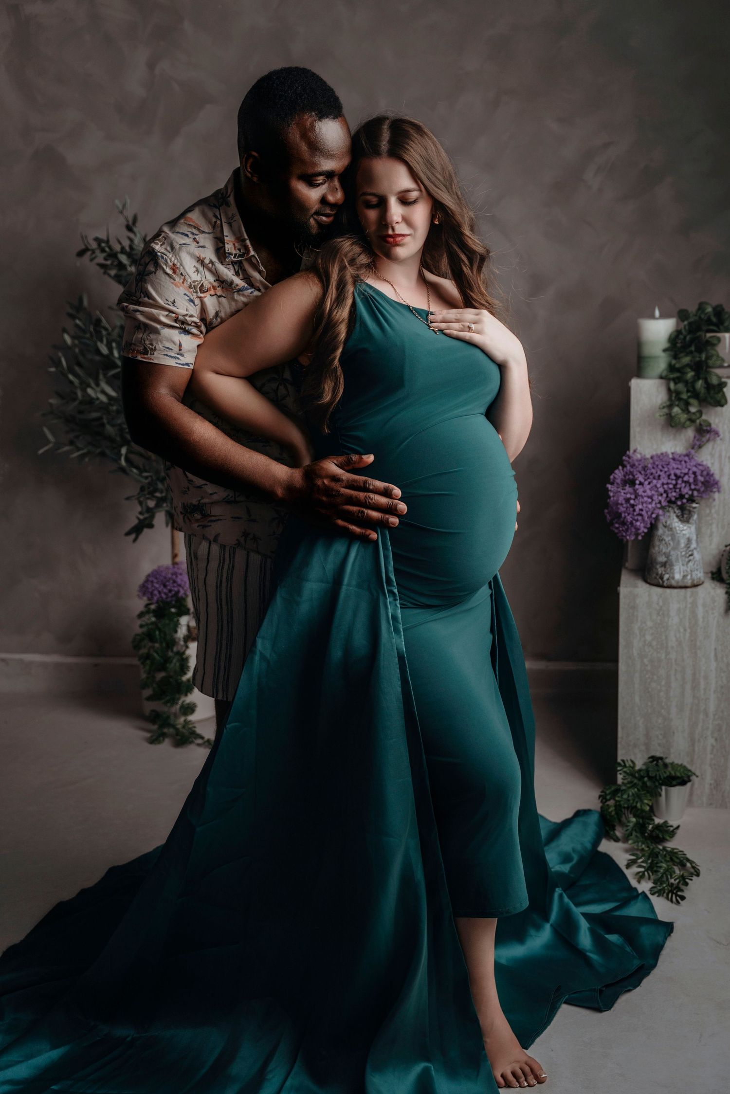 Couple in maternity photoshoot, woman in elegant teal gown, man embracing her from behind in studio setting.