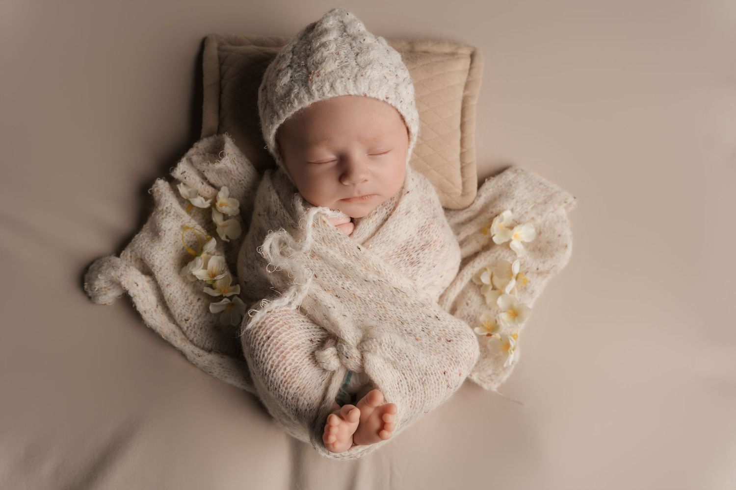 Peaceful newborn baby sleeping in cream knit outfit and hat on neutral beige blanket with tiny flowers.
