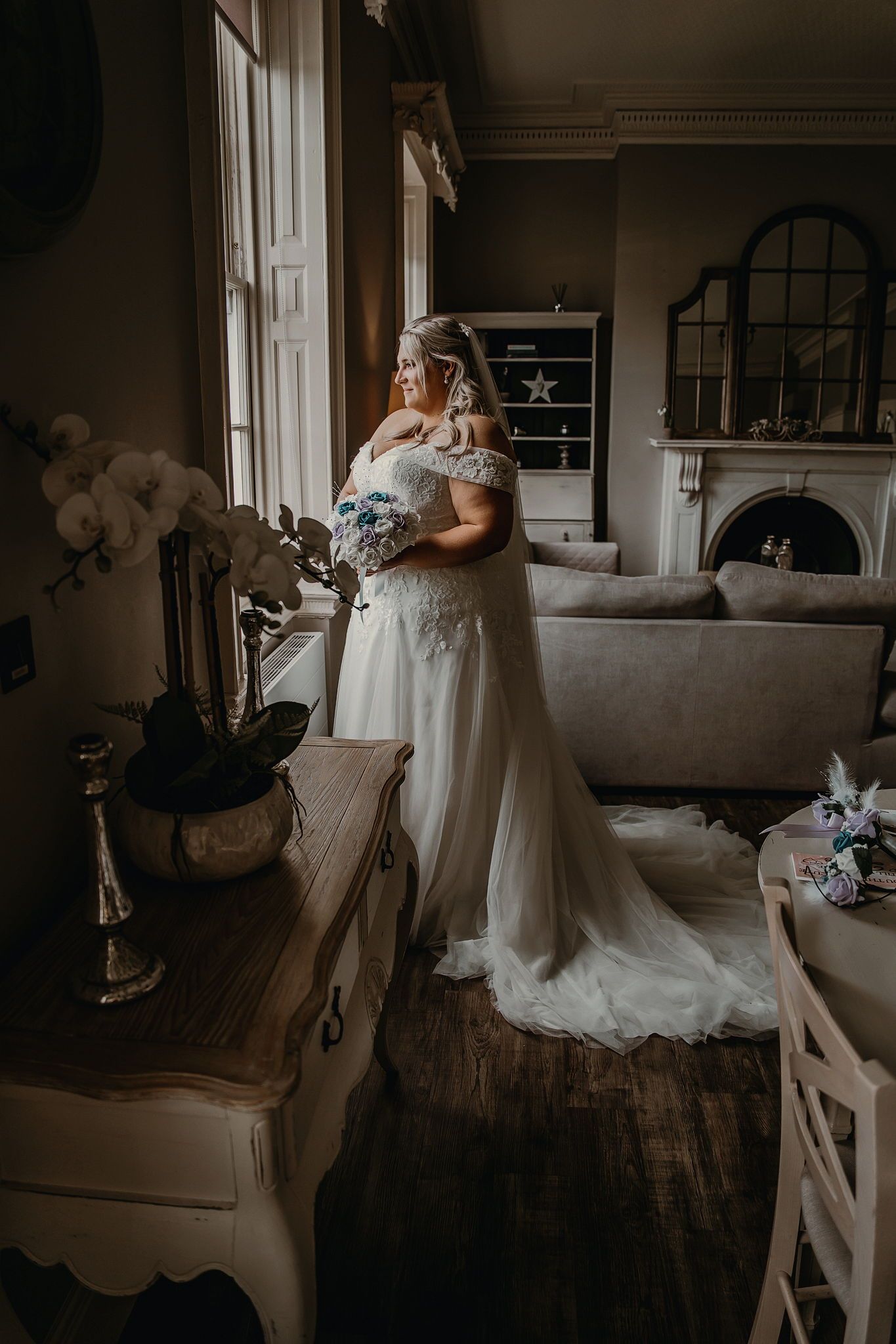 Bride in white off-shoulder gown with veil holds bouquet by window in elegant room with fireplace.