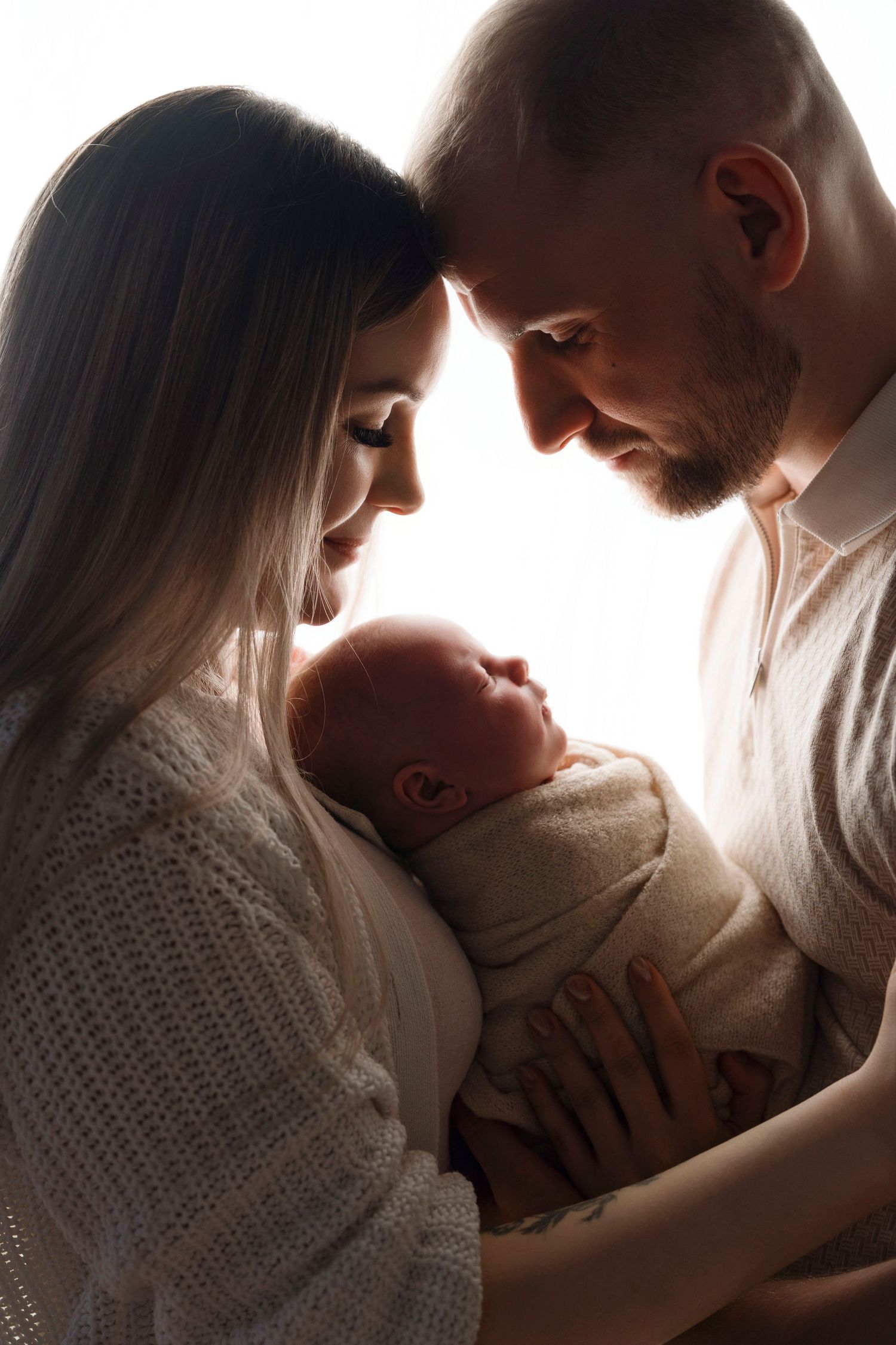 Silhouetted parents gazing lovingly at their swaddled newborn baby in soft backlit studio lighting.