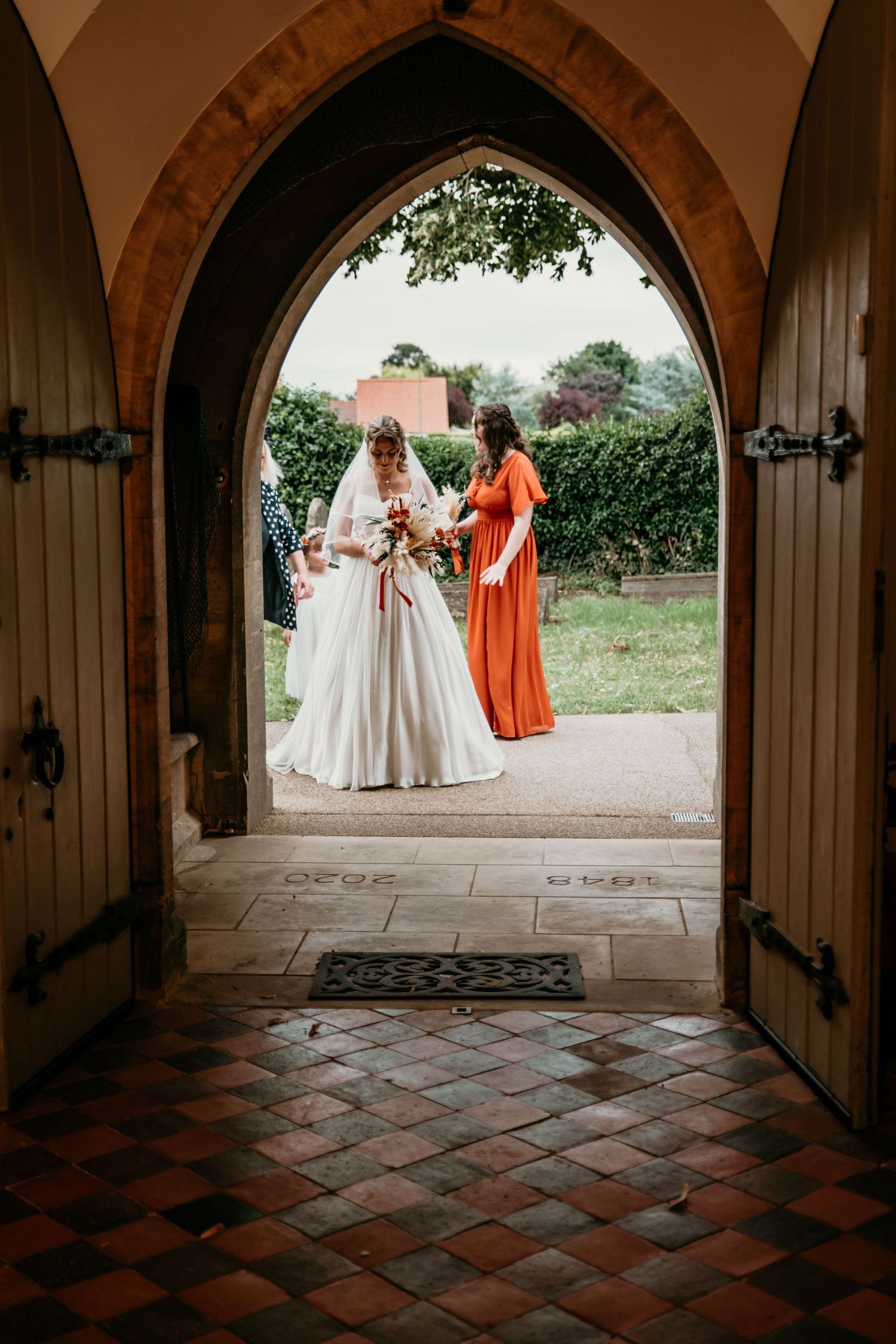 Bride in white gown with bridesmaid in orange dress framed by gothic church doorway.