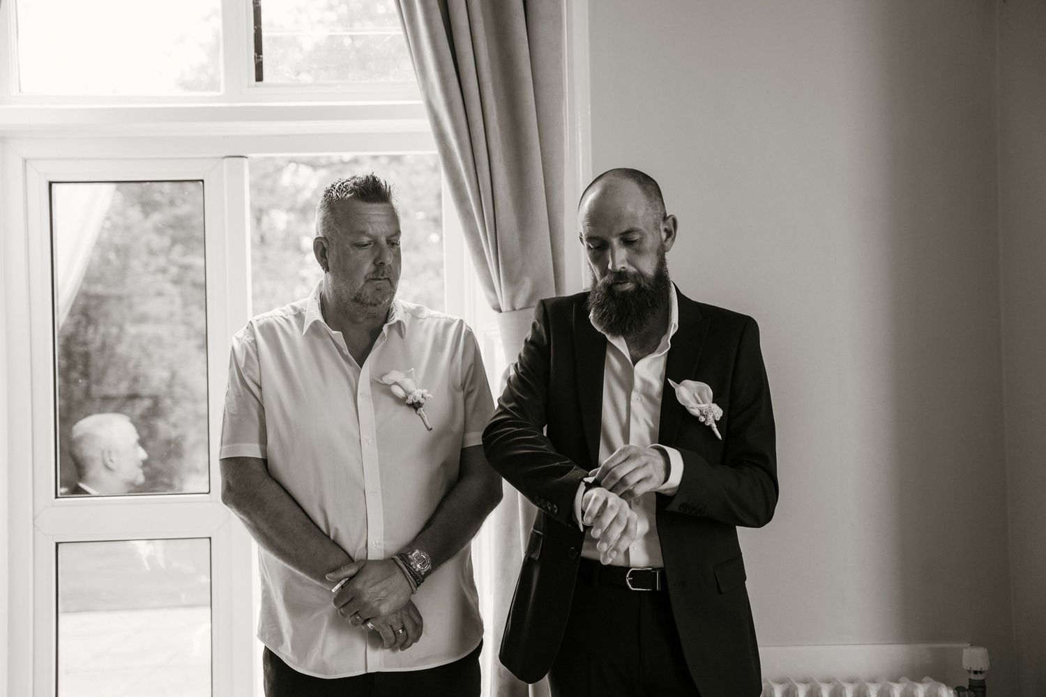 Groom adjusting cufflinks with best man beside him in black and white wedding preparation photo.