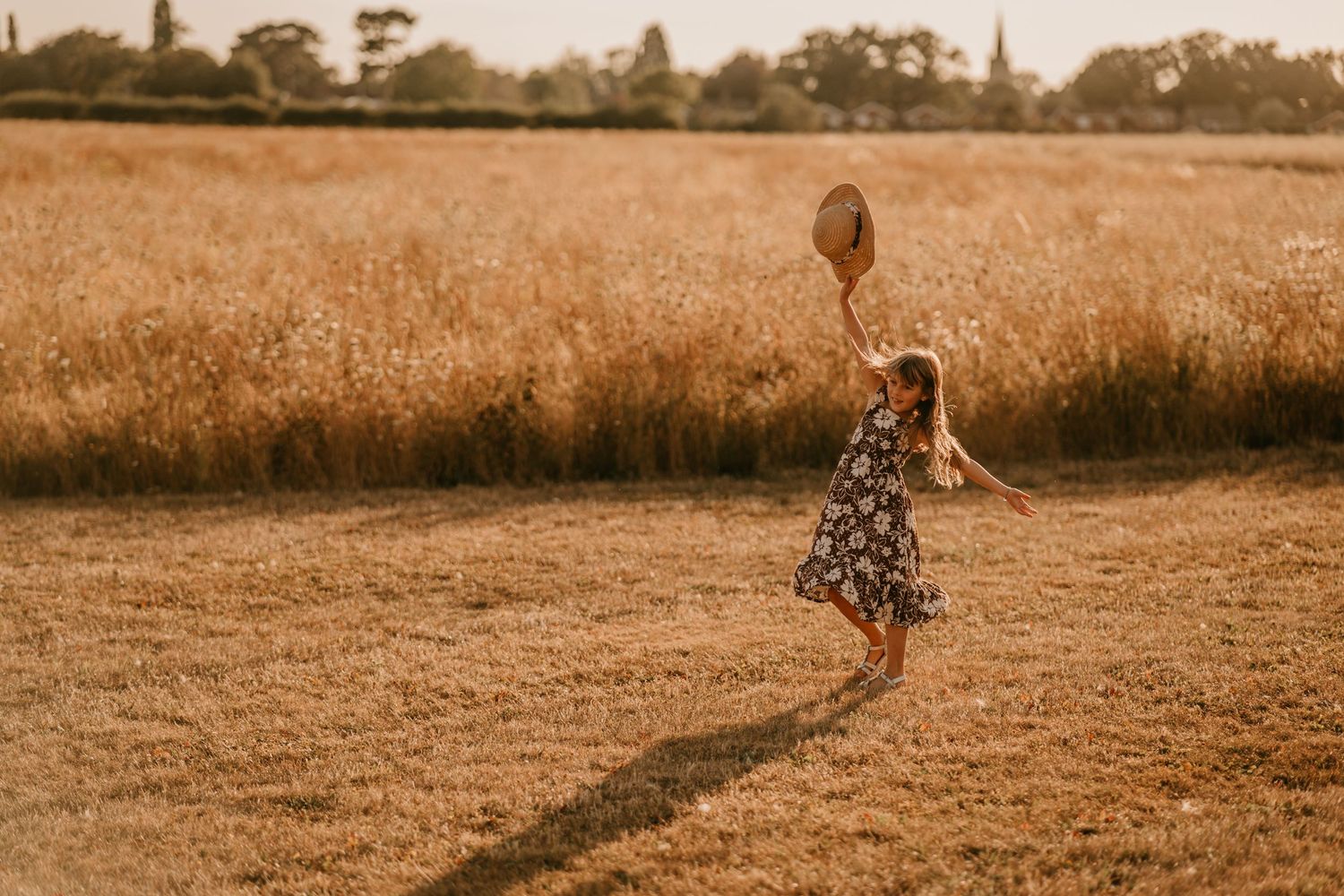 A young girl in a floral dress twirls joyfully in a golden wheat field, holding a straw hat aloft at sunset.