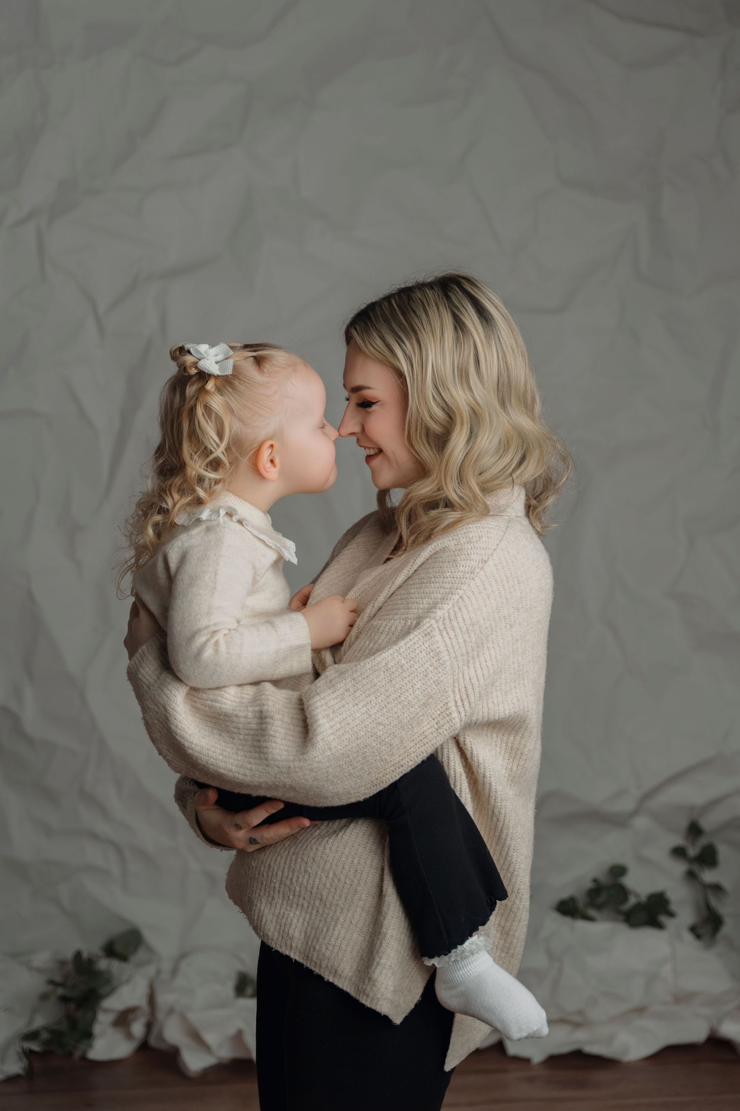 Blonde mother holding young daughter in matching cream knit sweaters, touching noses and smiling in studio.