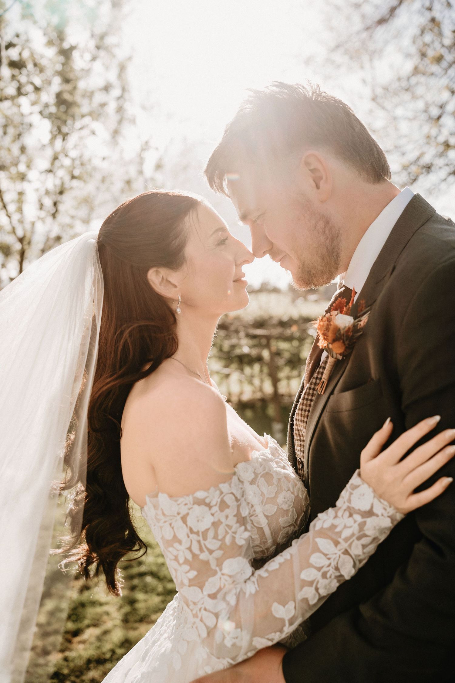 Bride in lace off-shoulder gown and groom in grey suit touching foreheads in golden sunlight outdoors.