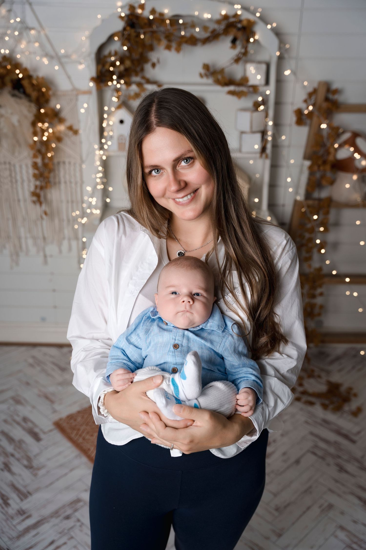 Smiling mother in a white shirt holding her newborn baby dressed in blue in a festive decorated room.