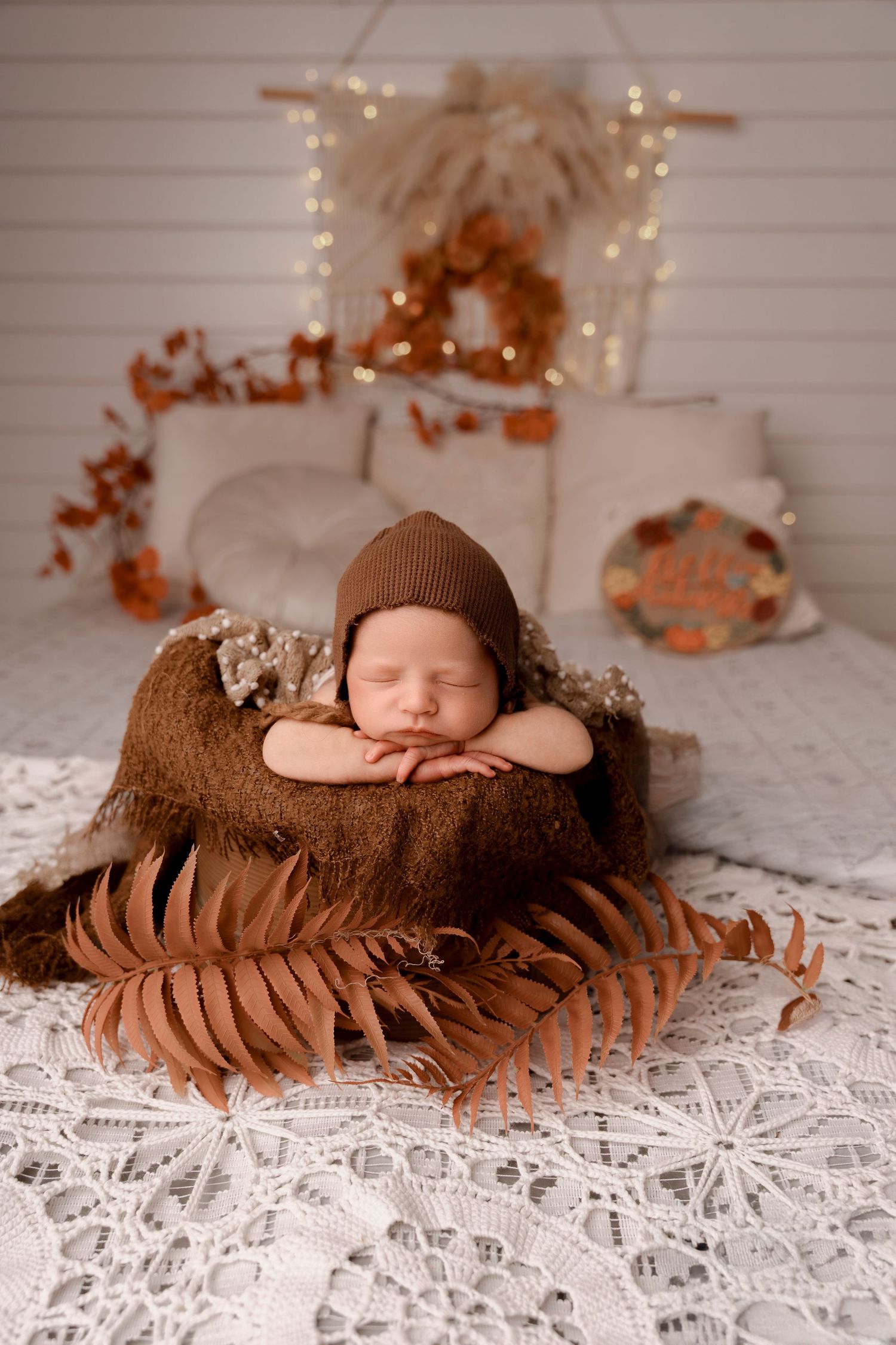 Sleeping newborn in brown knit bonnet posed in a wooden bowl surrounded by autumn fern leaves and bokeh lights.