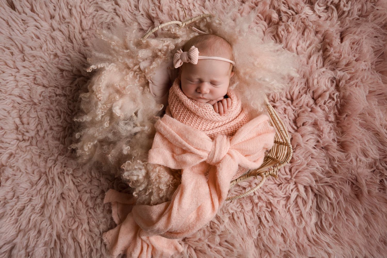 Sleeping newborn baby girl wrapped in peach knit swaddle with floral headband on fluffy pink fur backdrop.