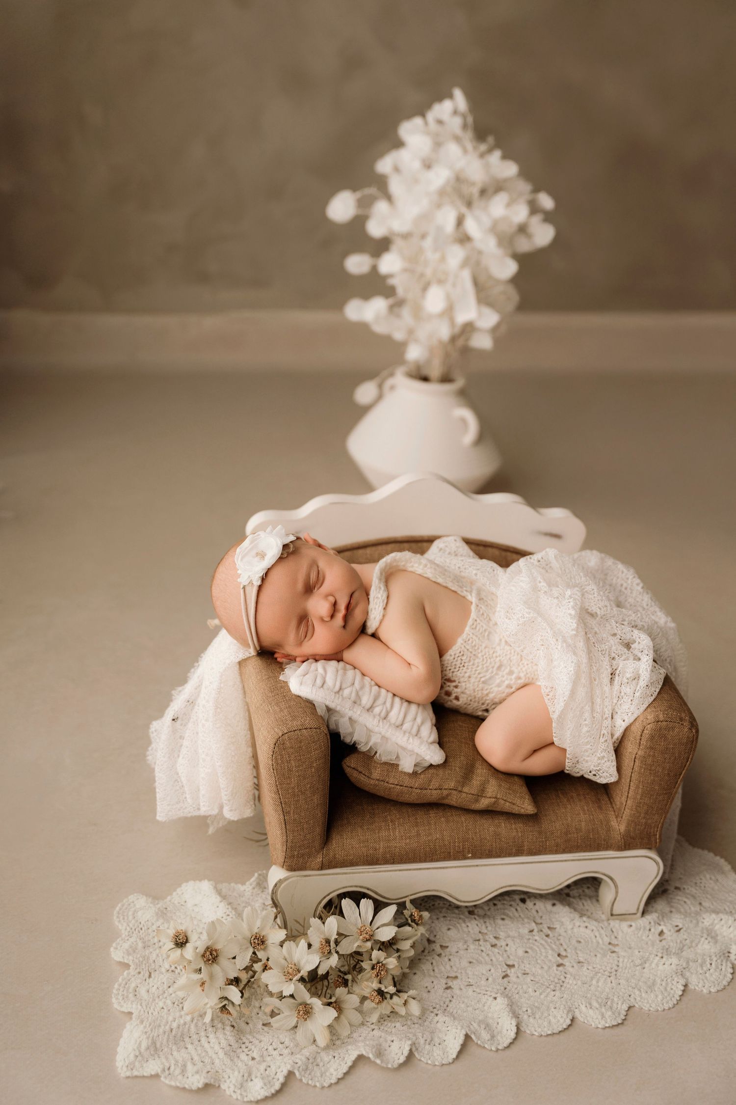 Smiling newborn baby in white lace dress sleeping on miniature sofa with white floral decor backdrop.