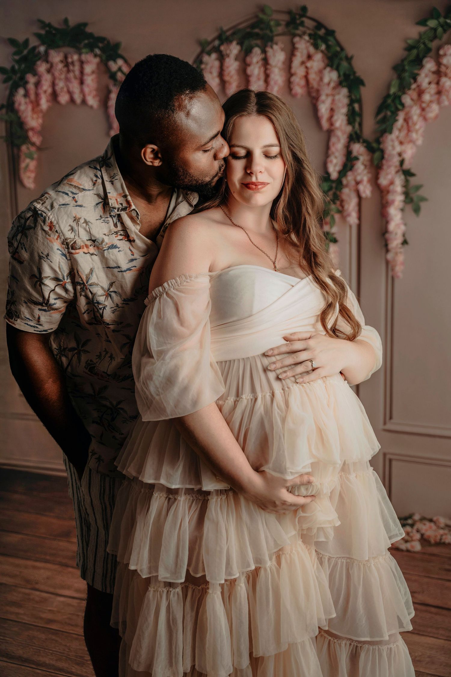 Interracial couple maternity photo shoot, man kissing pregnant woman in cream tulle gown with floral backdrop.
