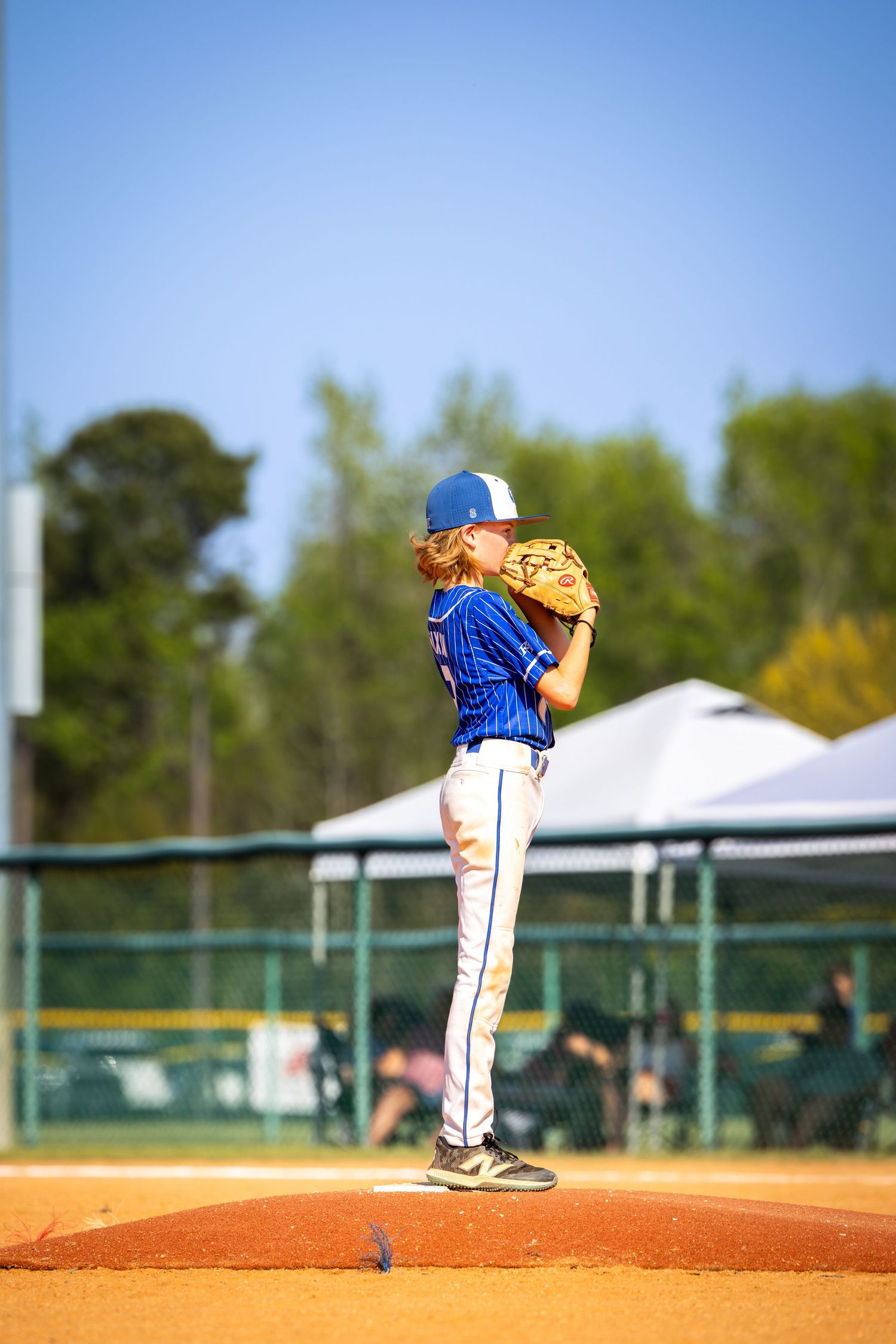 Young baseball pitcher in blue uniform winds up on the mound during a sunny youth league game.