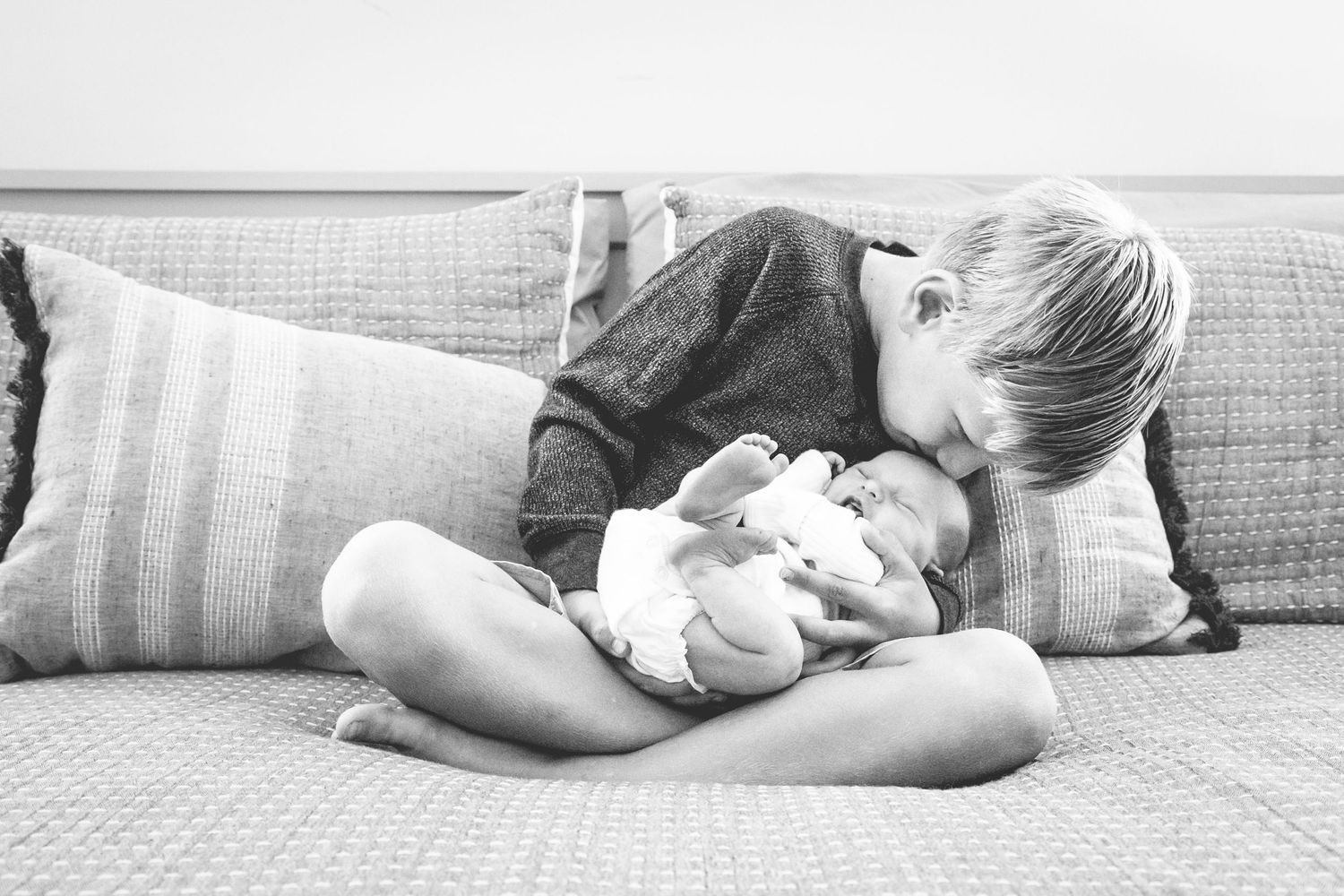 A child sits cross-legged on a couch with striped pillows, tenderly holding and looking at a baby in black and white.