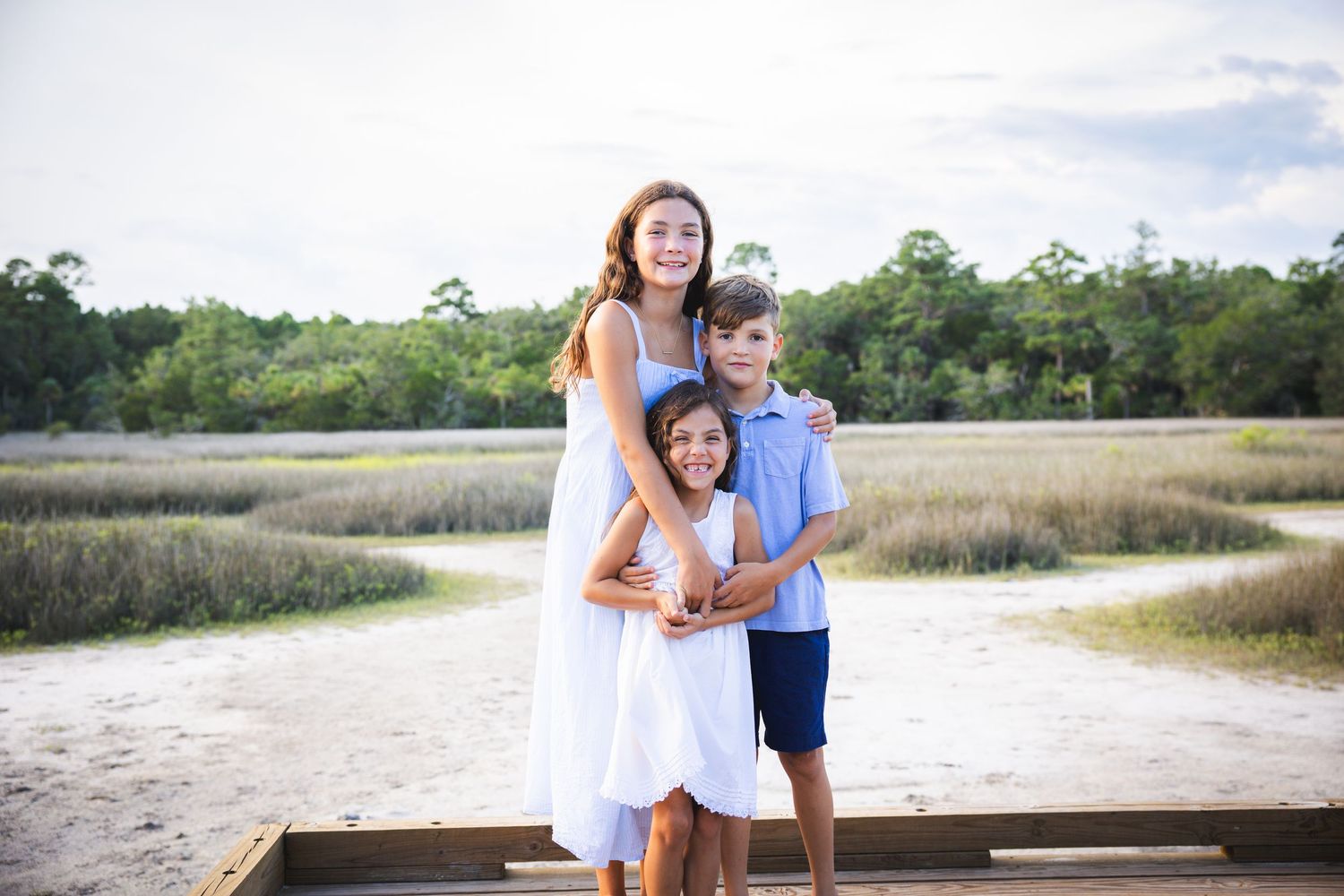 Three people embrace while standing on a wooden deck overlooking a scenic coastal marsh landscape on a sunny day.