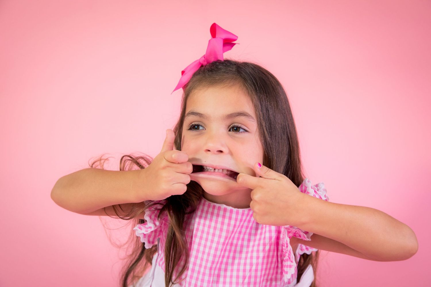 Young child in pink gingham dress and bow against pink background covering mouth with both hands in playful gesture.