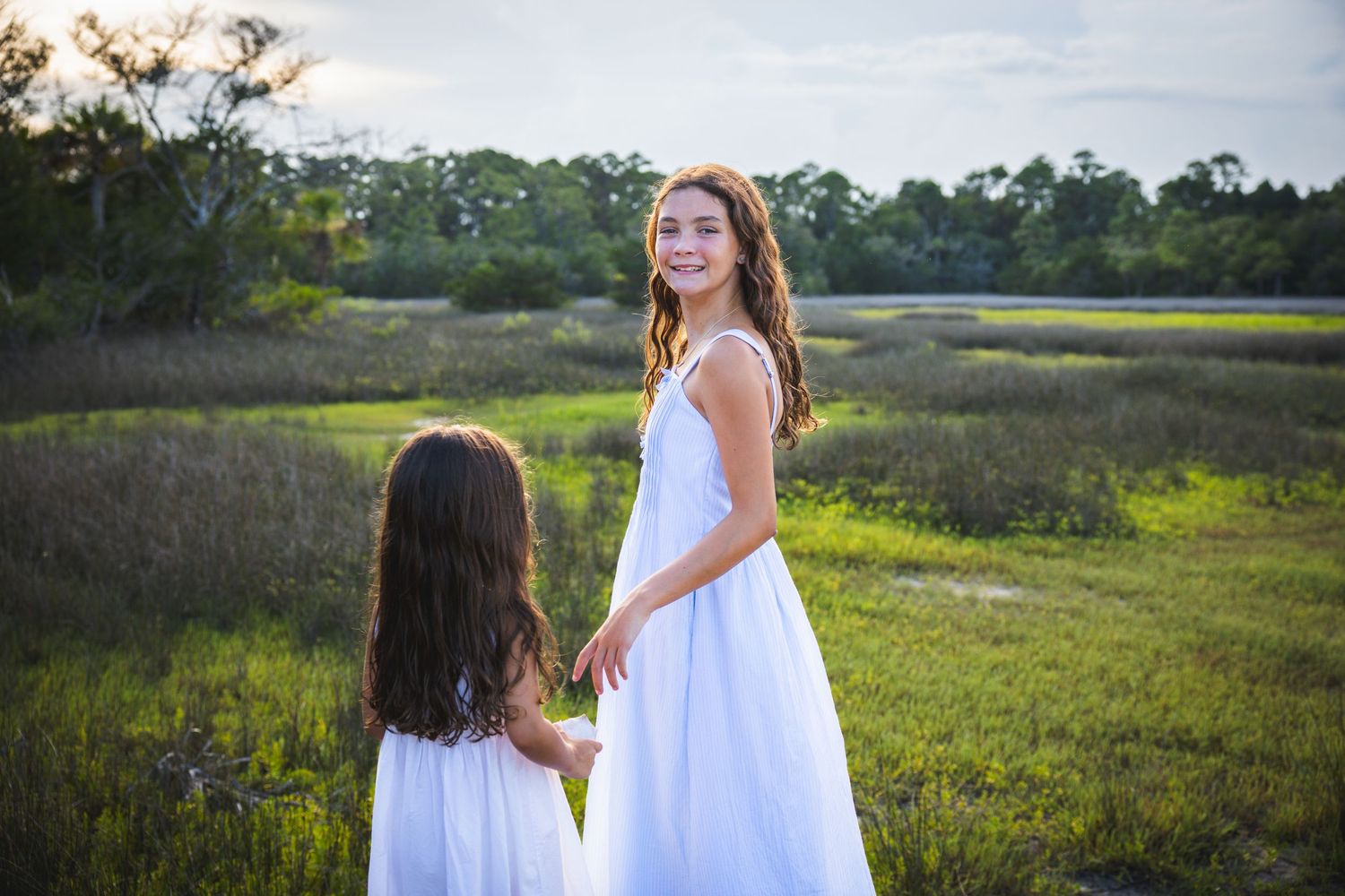 Two figures in flowing white dresses stand together in a sunlit meadow during golden hour.