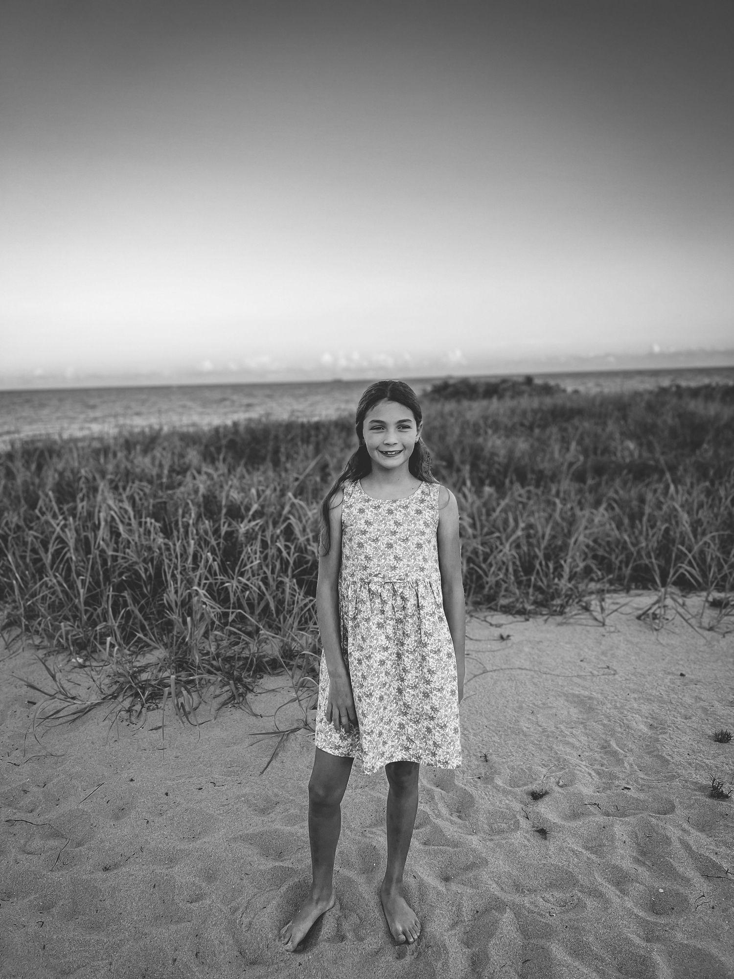 A black and white photo of someone in a floral dress standing on sand dunes against a hazy sky.