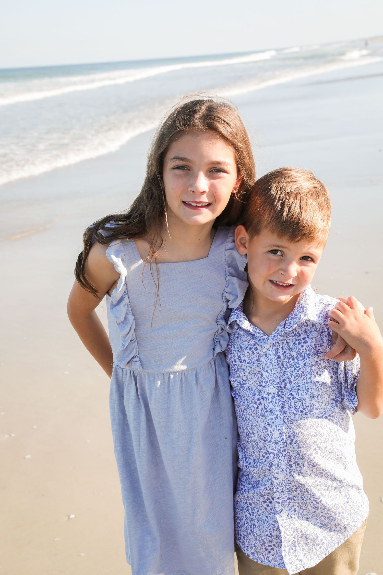 Two children stand together smiling on a sunny beach wearing light blue and floral patterned clothing.