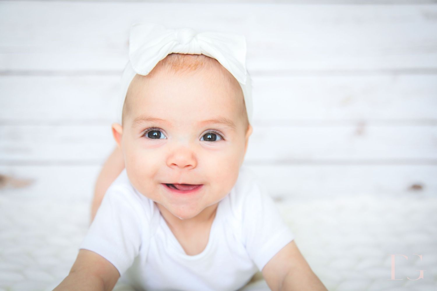 Smiling baby wearing white clothing lying on soft white surface.