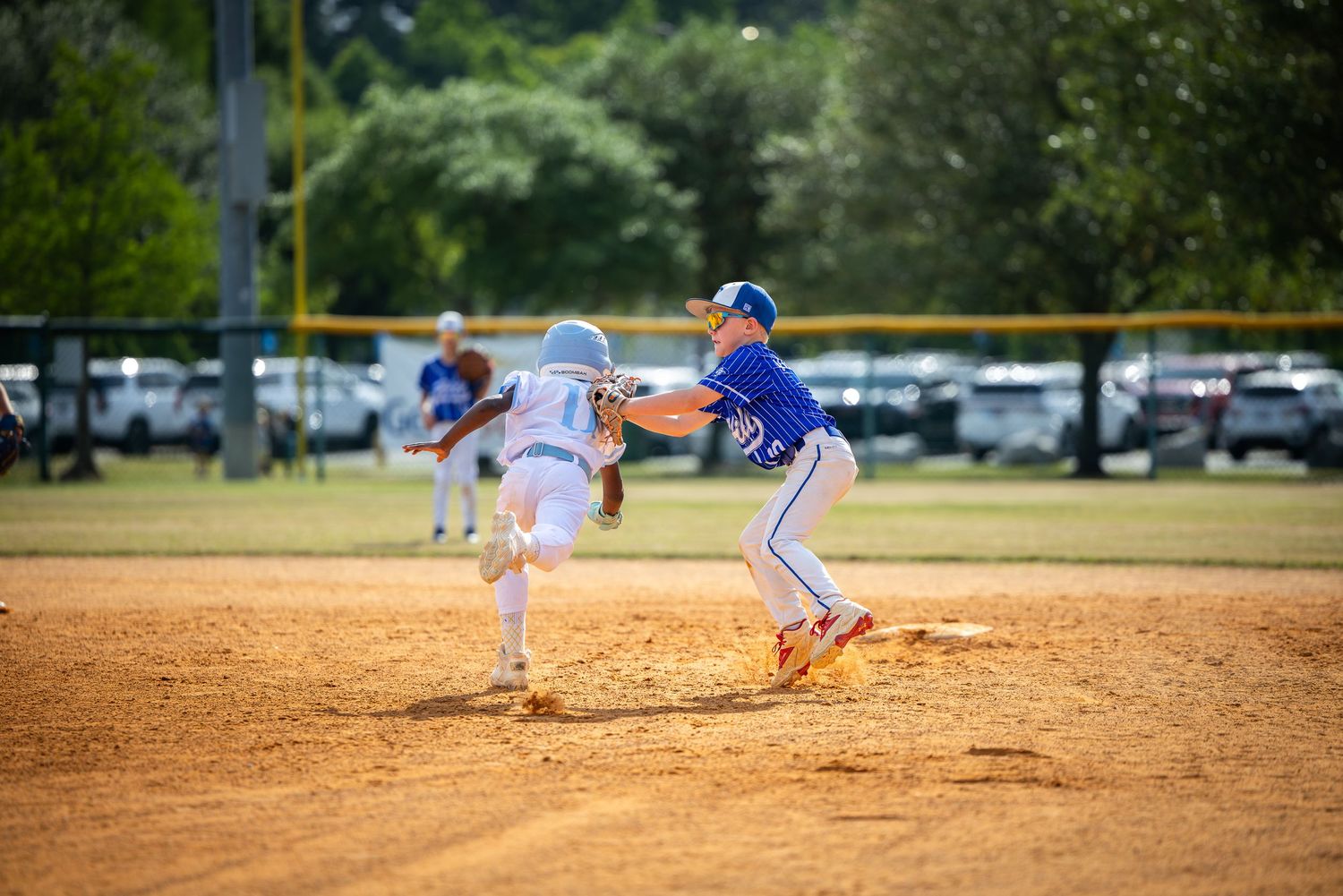Young baseball players compete in a youth league game, with a fielder catching the ball at base on a sunny day.