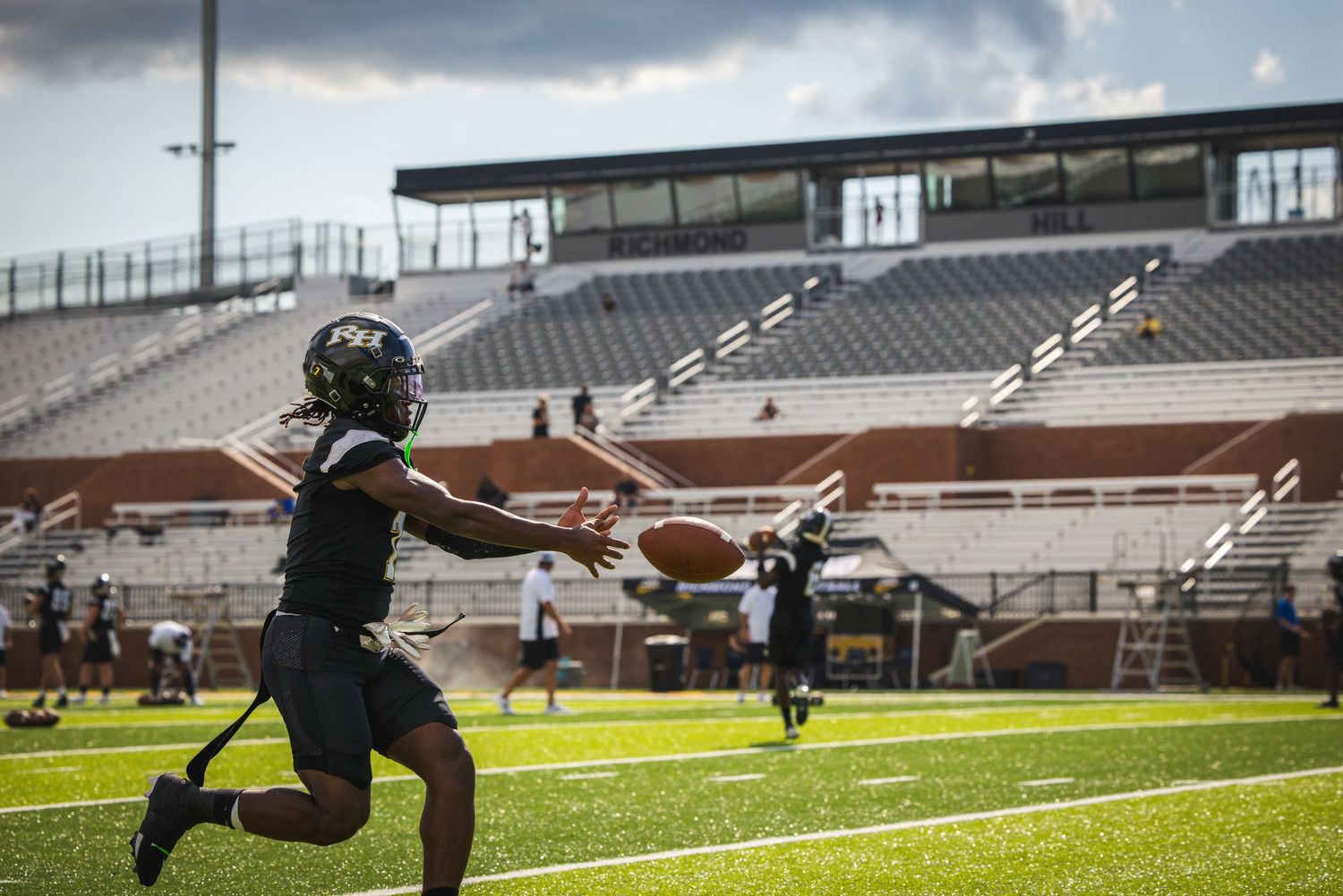 Football player catching a pass on a green field during practice at a stadium with empty bleachers.