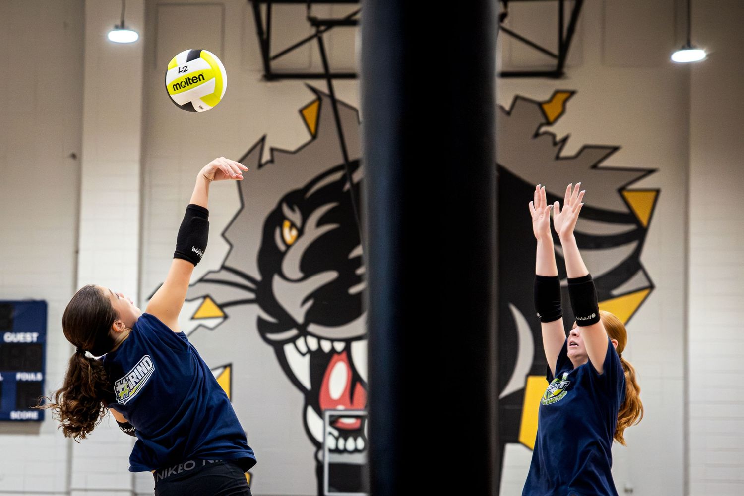 Girls playing volleyball in a gym, one spiking a yellow ball while another attempts to block at the net.