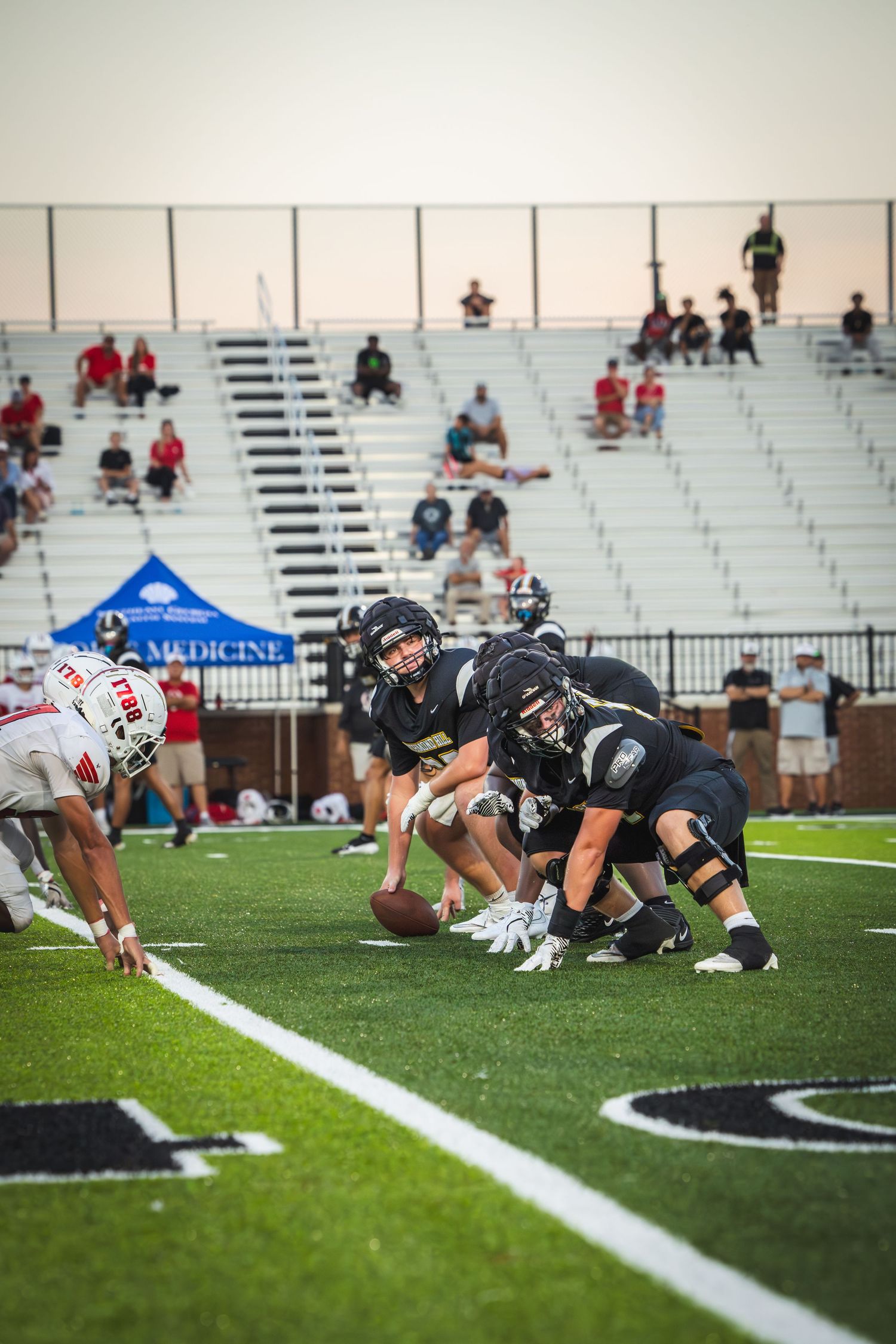 Football players line up at scrimmage during a game with spectators watching from bleachers in the background.