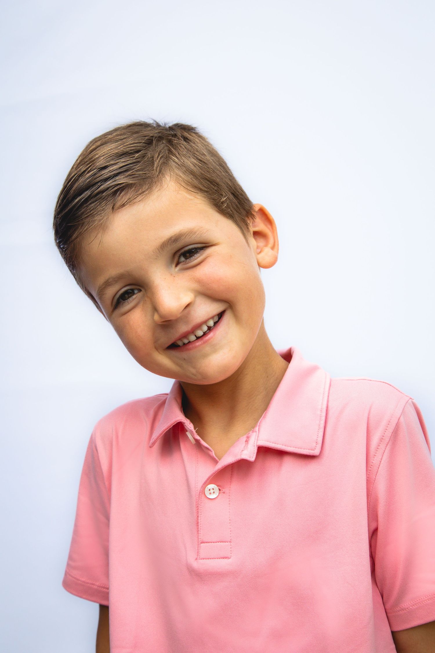 A child wearing a pink polo shirt tilts their head and smiles warmly at the camera against a white background.