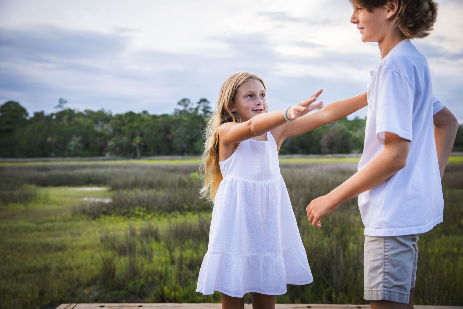 Children playing and reaching for each other on a wooden walkway overlooking coastal marshland.