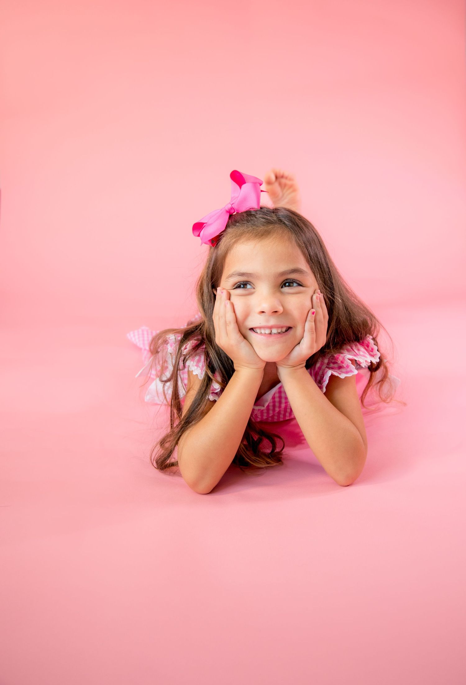 Sweet child in pink outfit laying on pink backdrop with hands under chin and bow hair accessory.