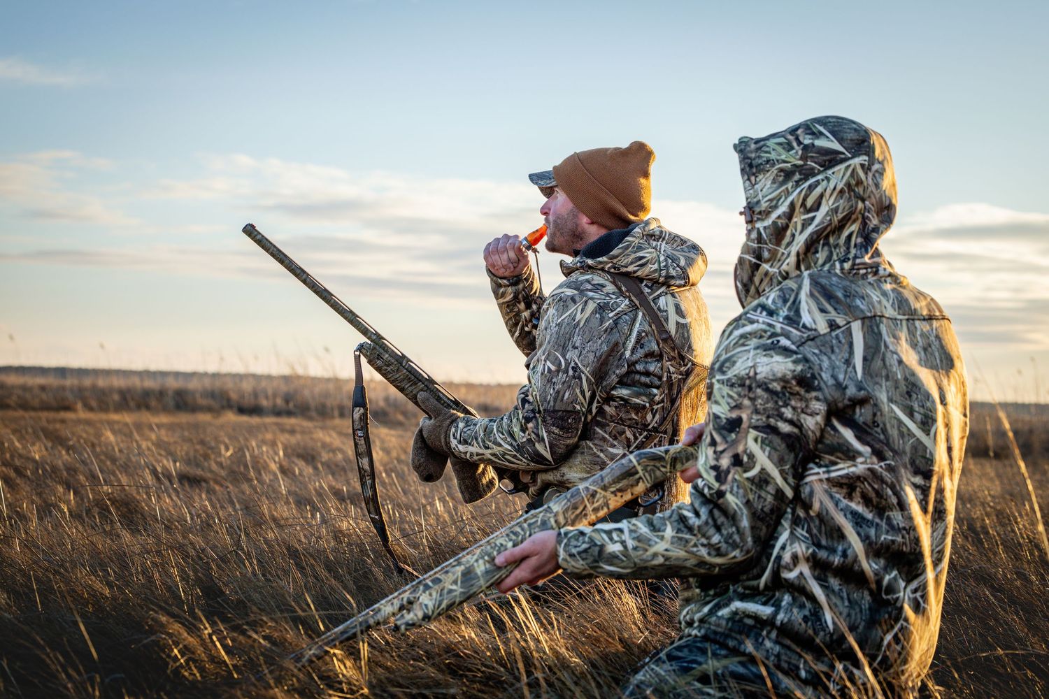 Two hunters in camouflage gear sitting in tall grass at sunset with rifles during hunting season.
