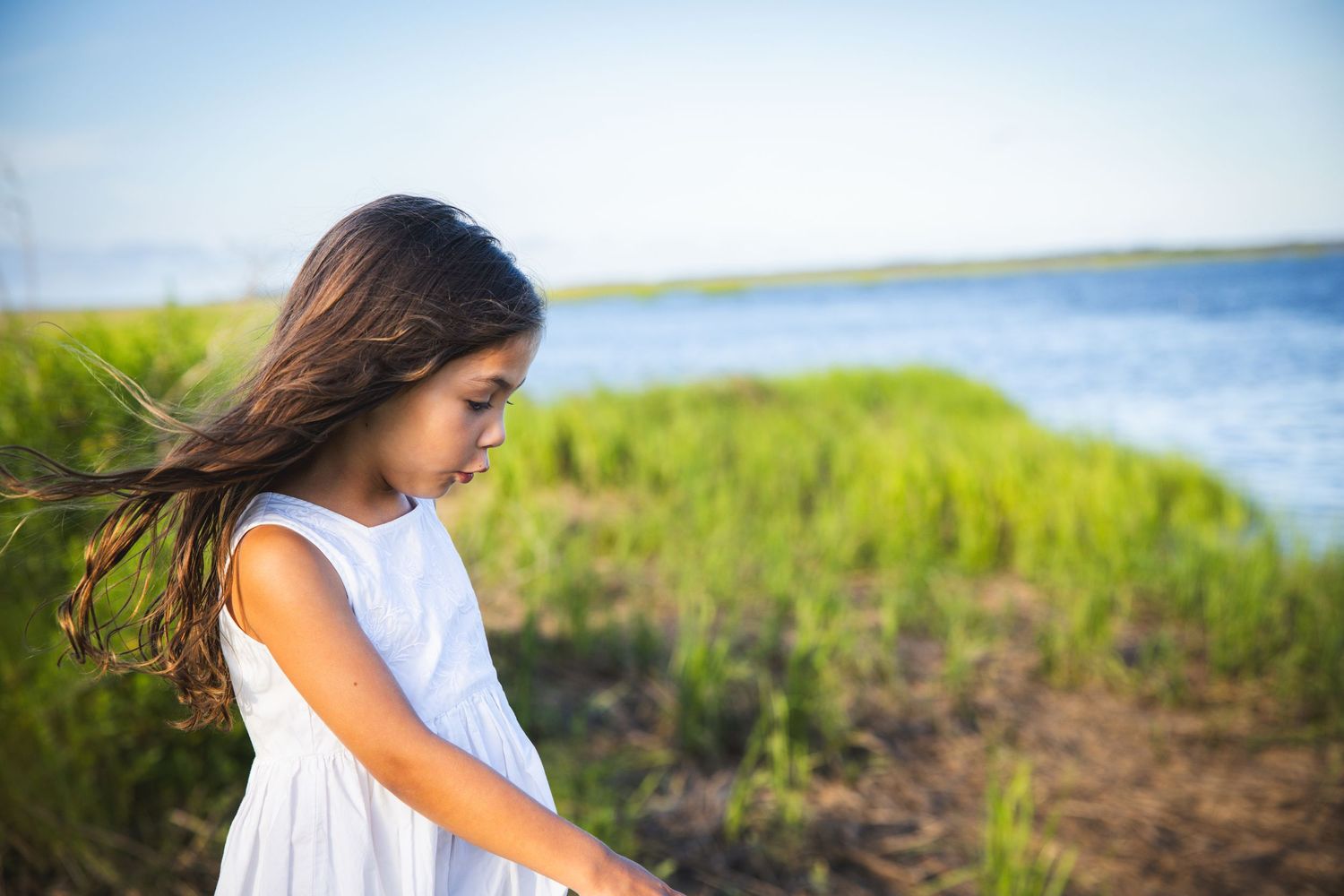 Young child in white dress looking down thoughtfully near coastal waters and marsh grass.