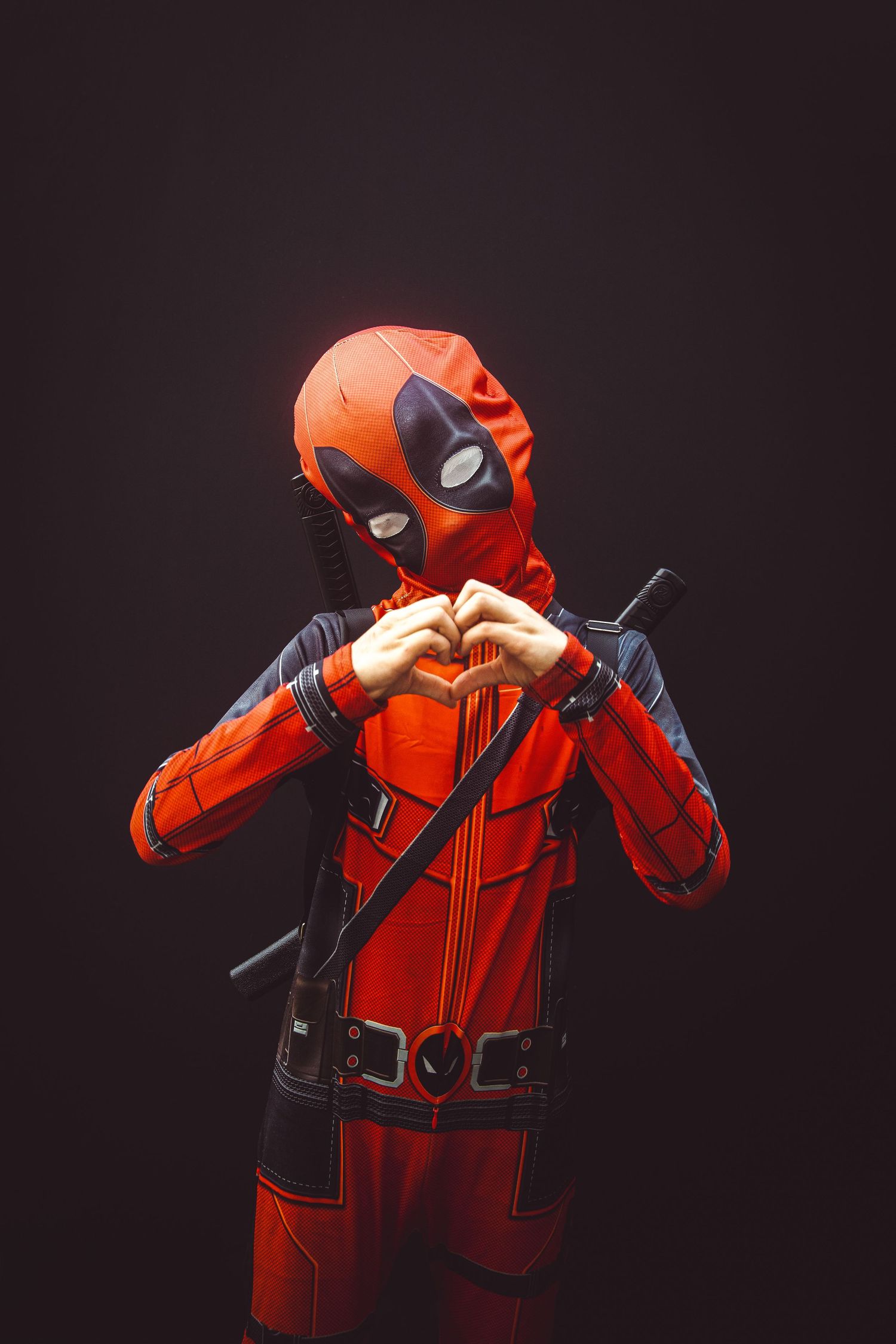 Cosplayer in a red and black Deadpool costume makes a heart shape gesture against dark background.