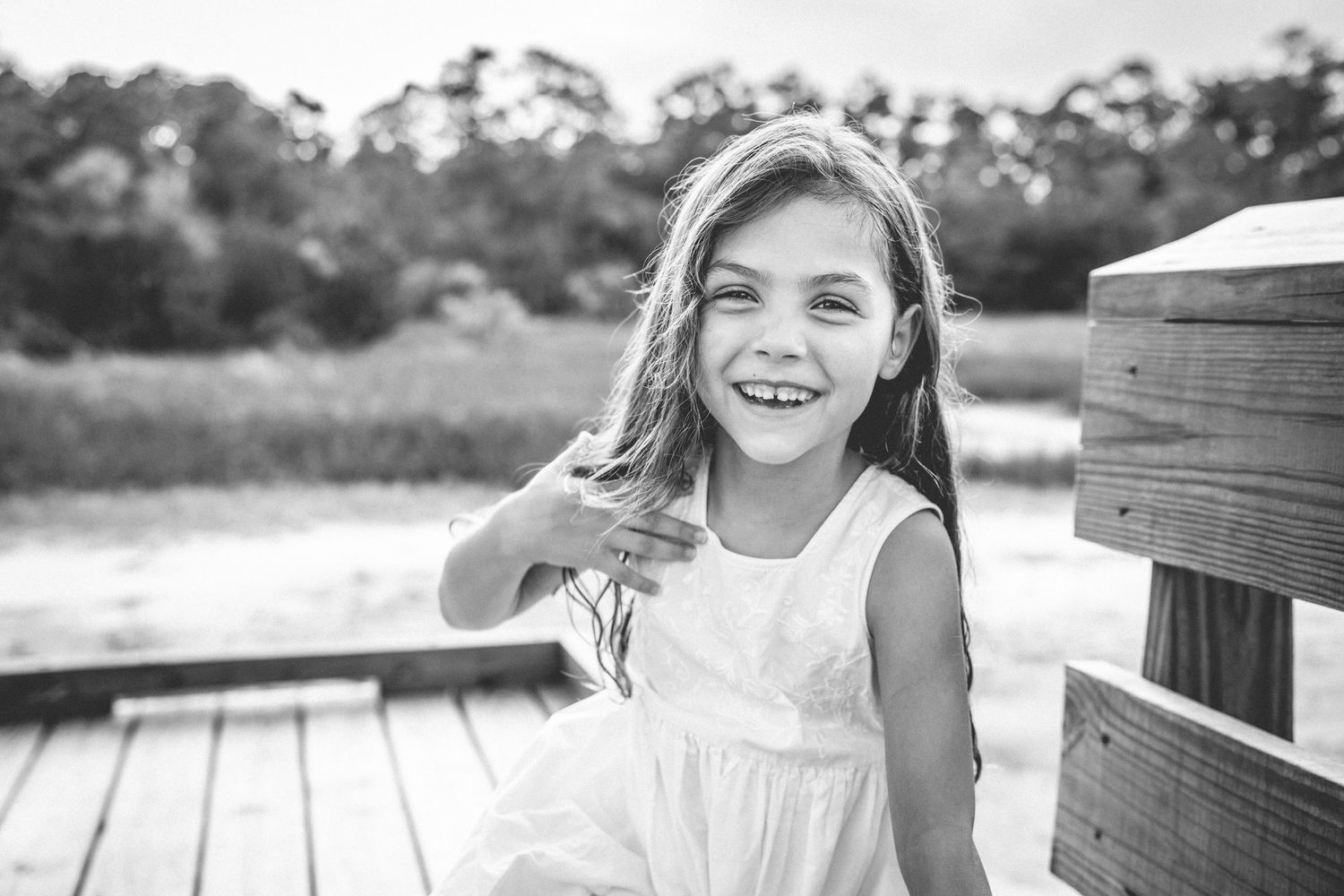 A child in a flowing white dress smiles while standing on a wooden deck with trees in the background.