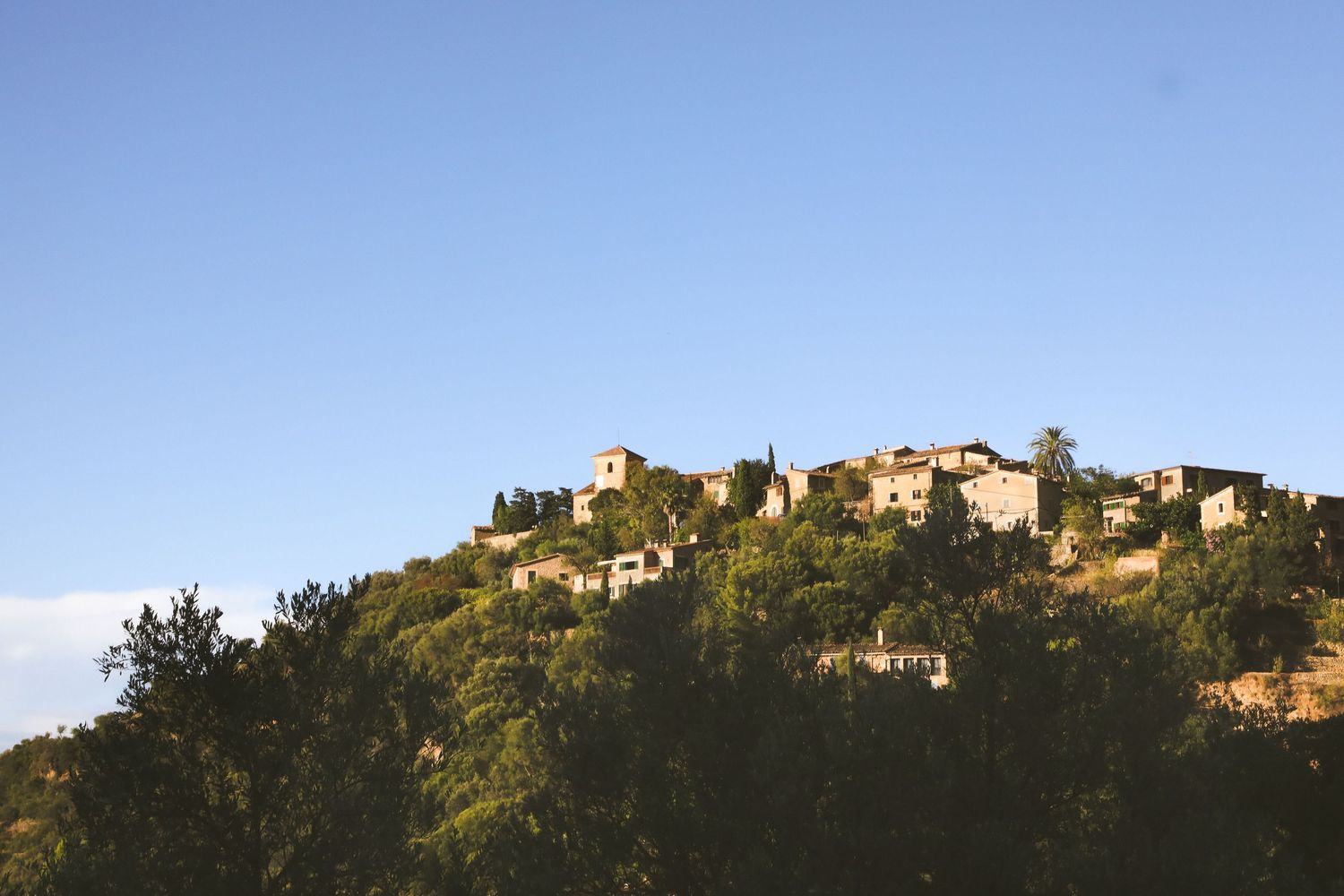 A hillside Mediterranean village with stone buildings perched atop a green slope against a bright blue sky.