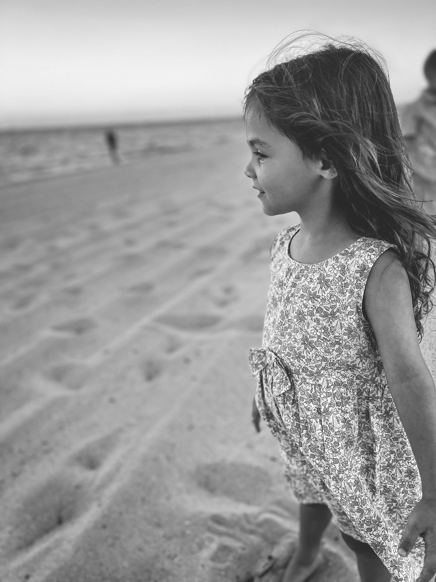 A young girl in a floral dress plays on a sandy beach, shown in a series of black and white candid moments.