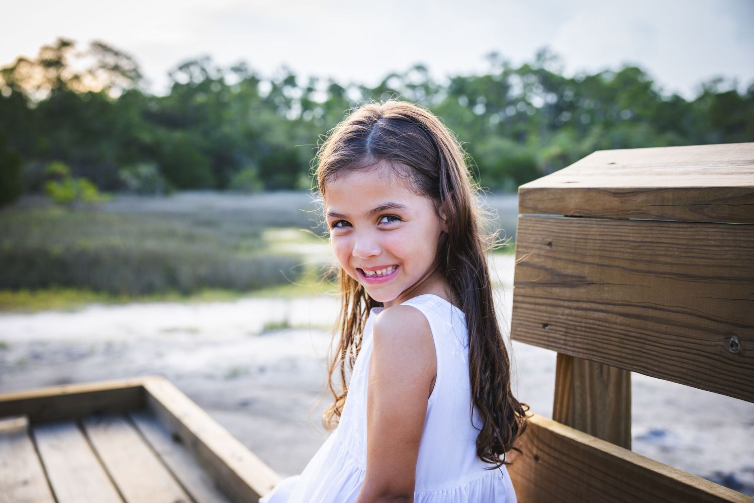 A person wearing a white dress walks on a wooden boardwalk pathway at a beach during sunset.