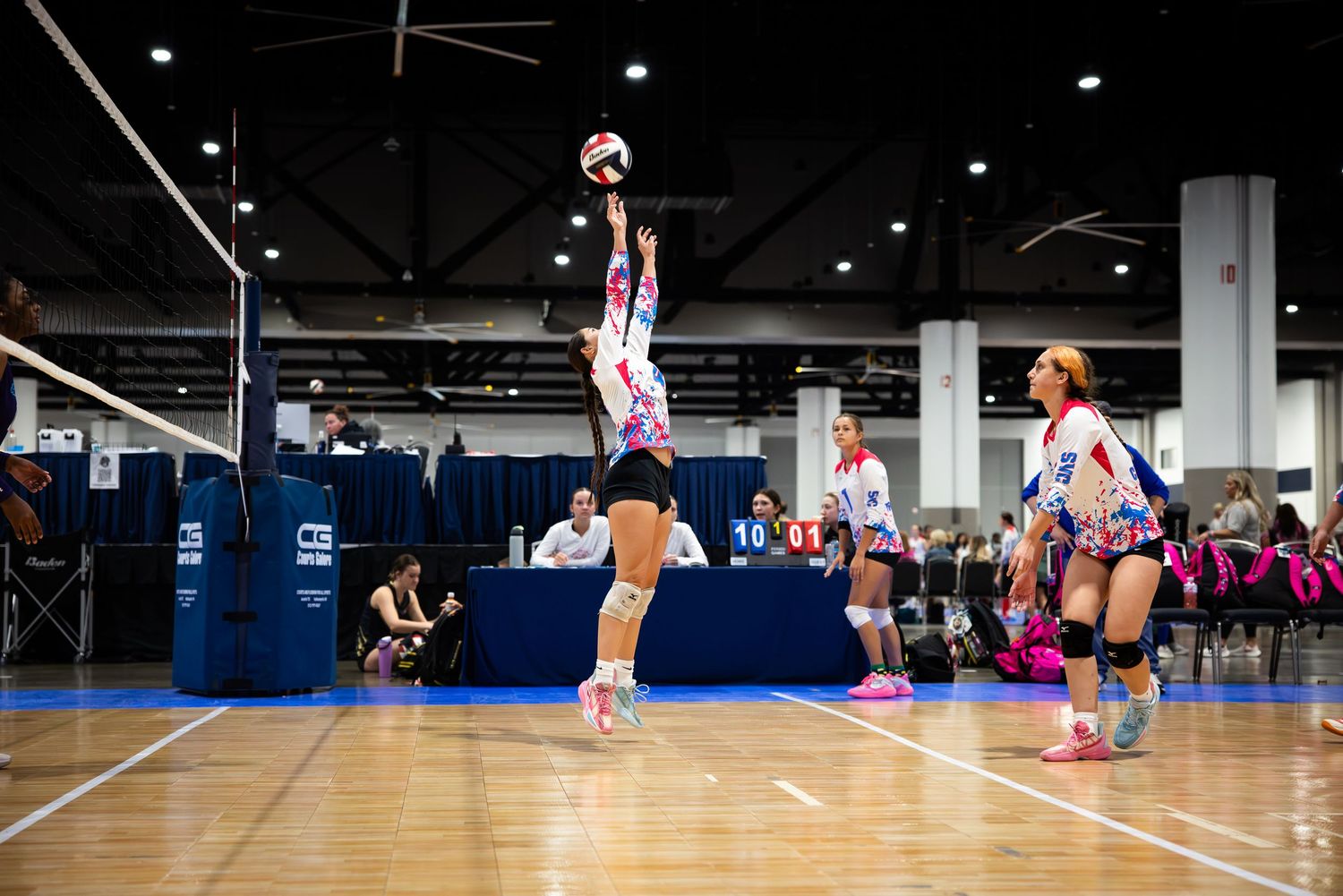 Female volleyball player sets the ball during an indoor tournament match in a large convention center gymnasium.