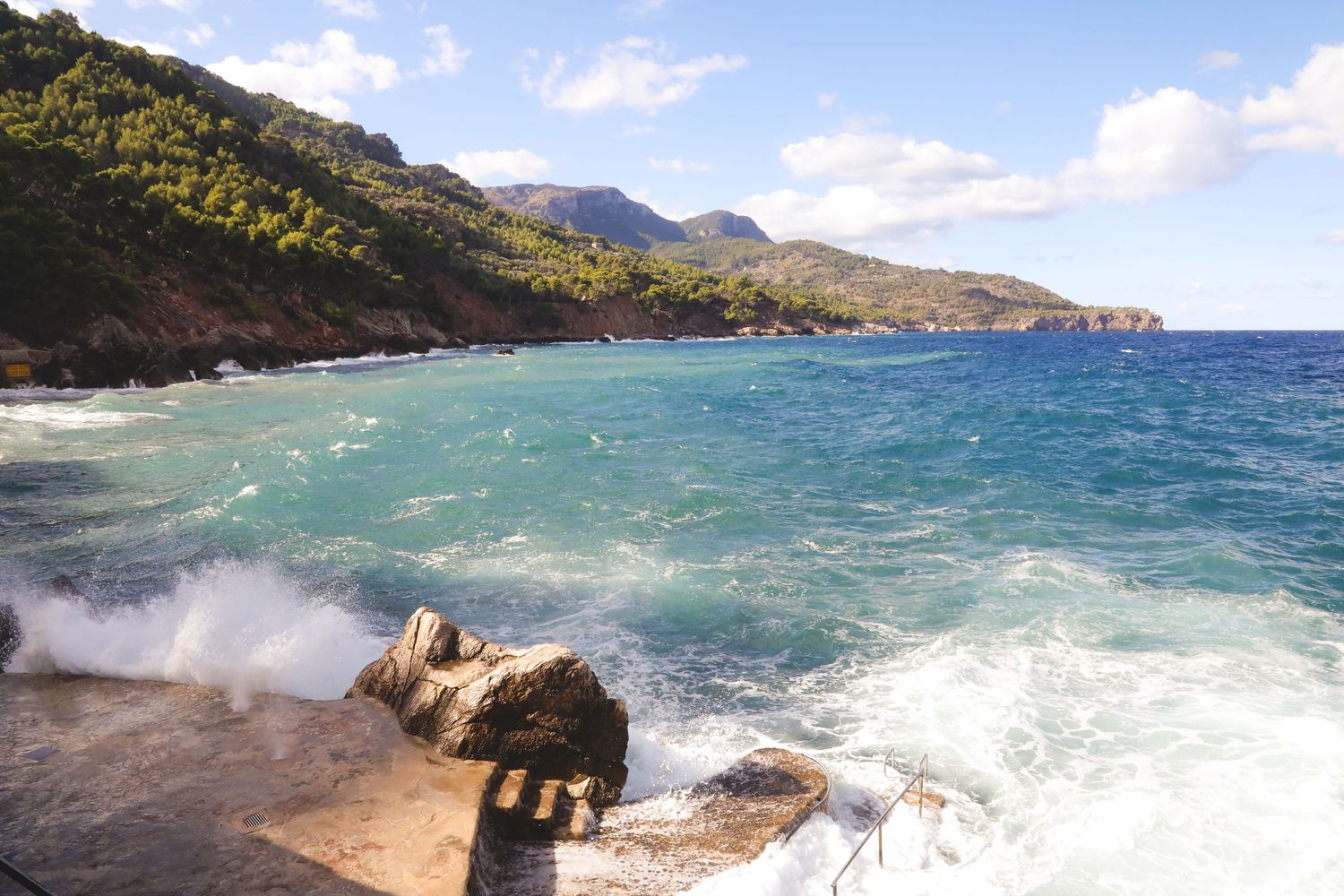 Turquoise waves crash against rocky coastline with mountains and blue sky in the background.